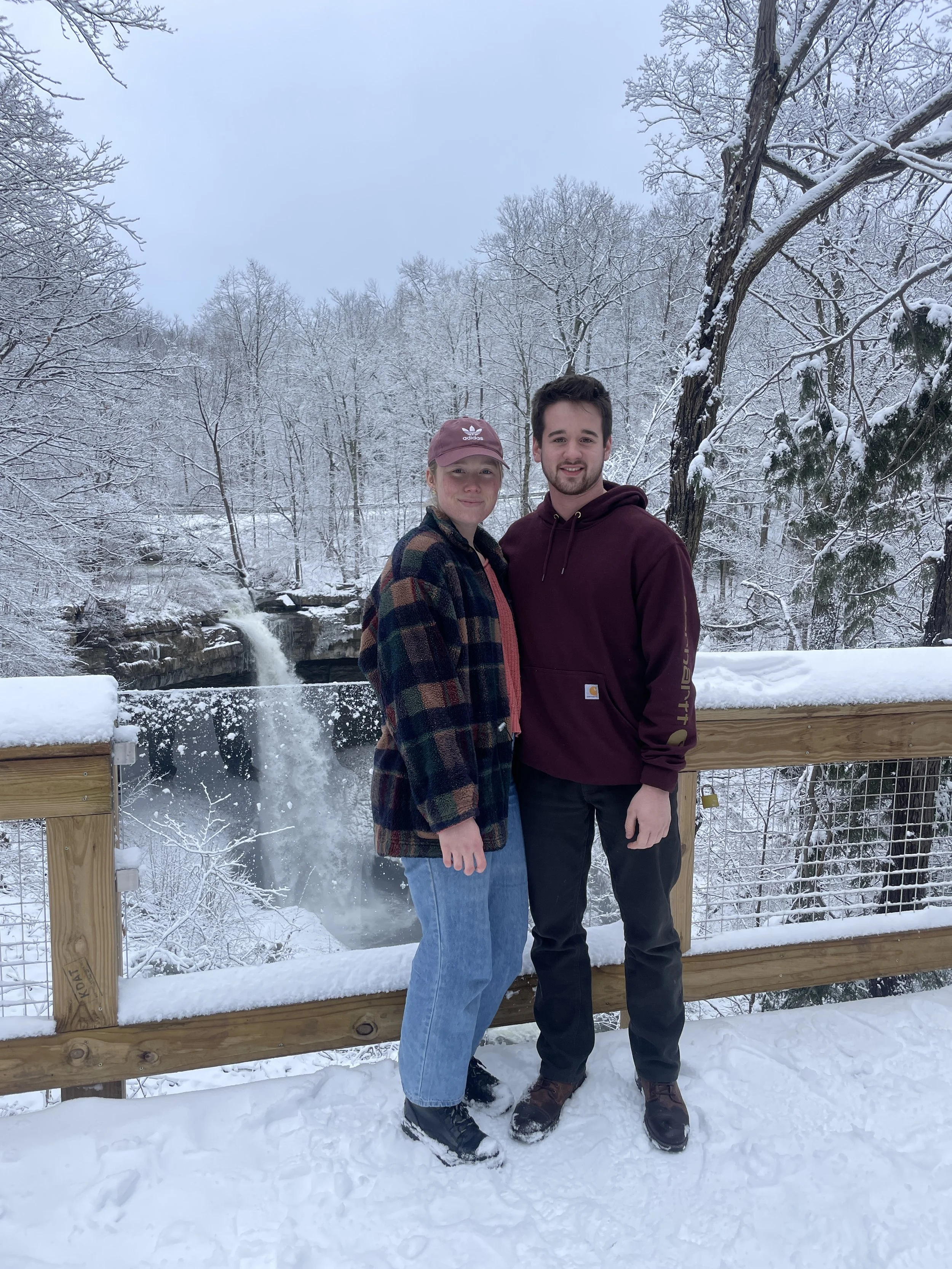 Two young adults, a woman and a man, standing on a snow-covered wooden bridge in a winter landscape with bare trees and a small waterfall in the background.