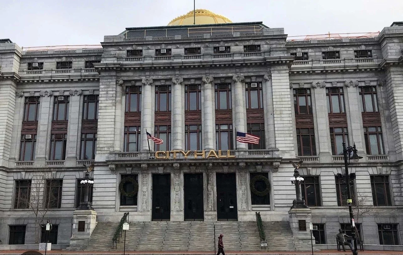 Facade of a historic city hall building with gray stone exterior, tall windows, and American flags, with steps leading to the entrance.