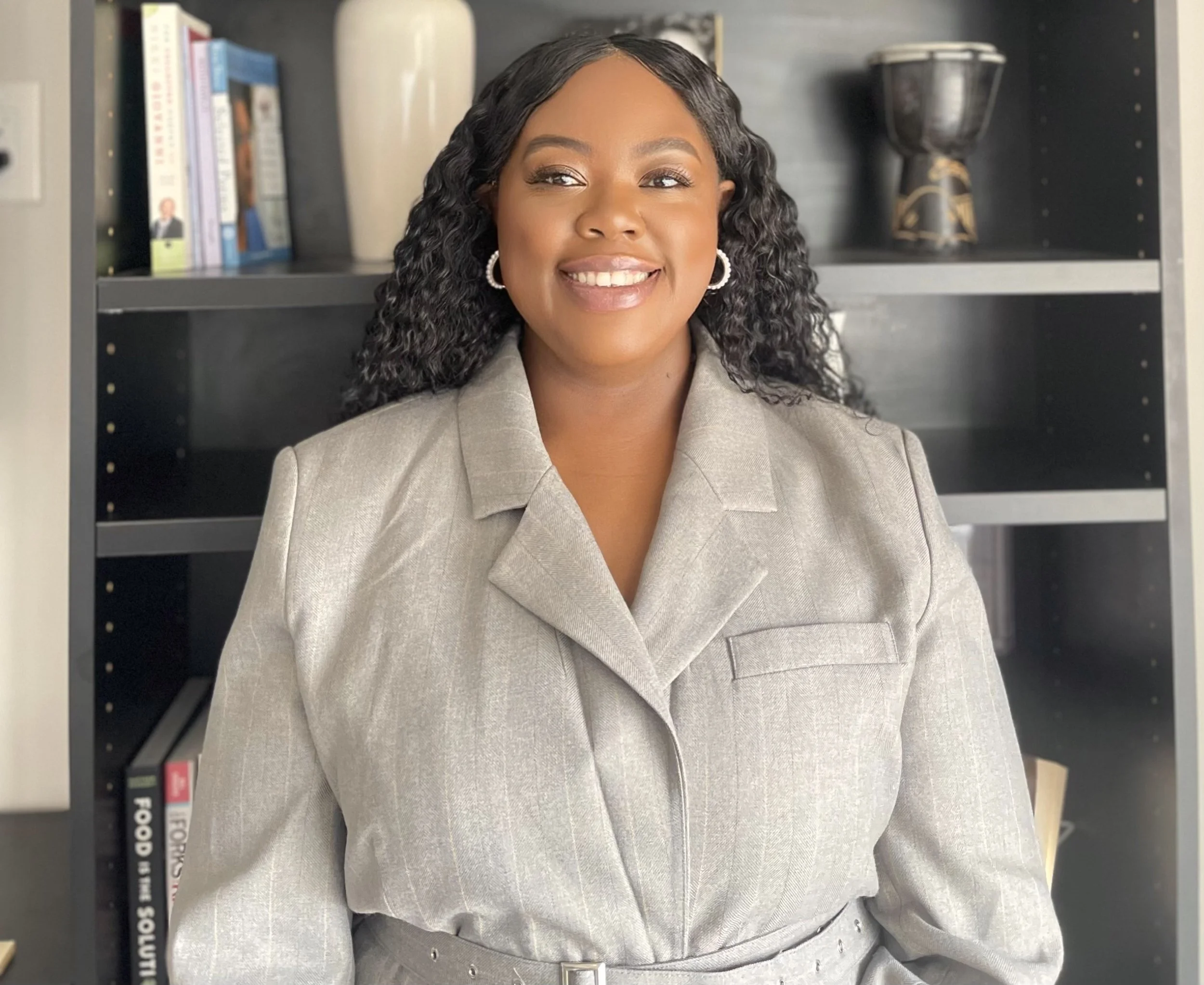 A woman with black curly hair wearing a beige blazer and pearl earrings smiling in front of a black bookshelf with books, a beige vase, and a decorative black and gold item.