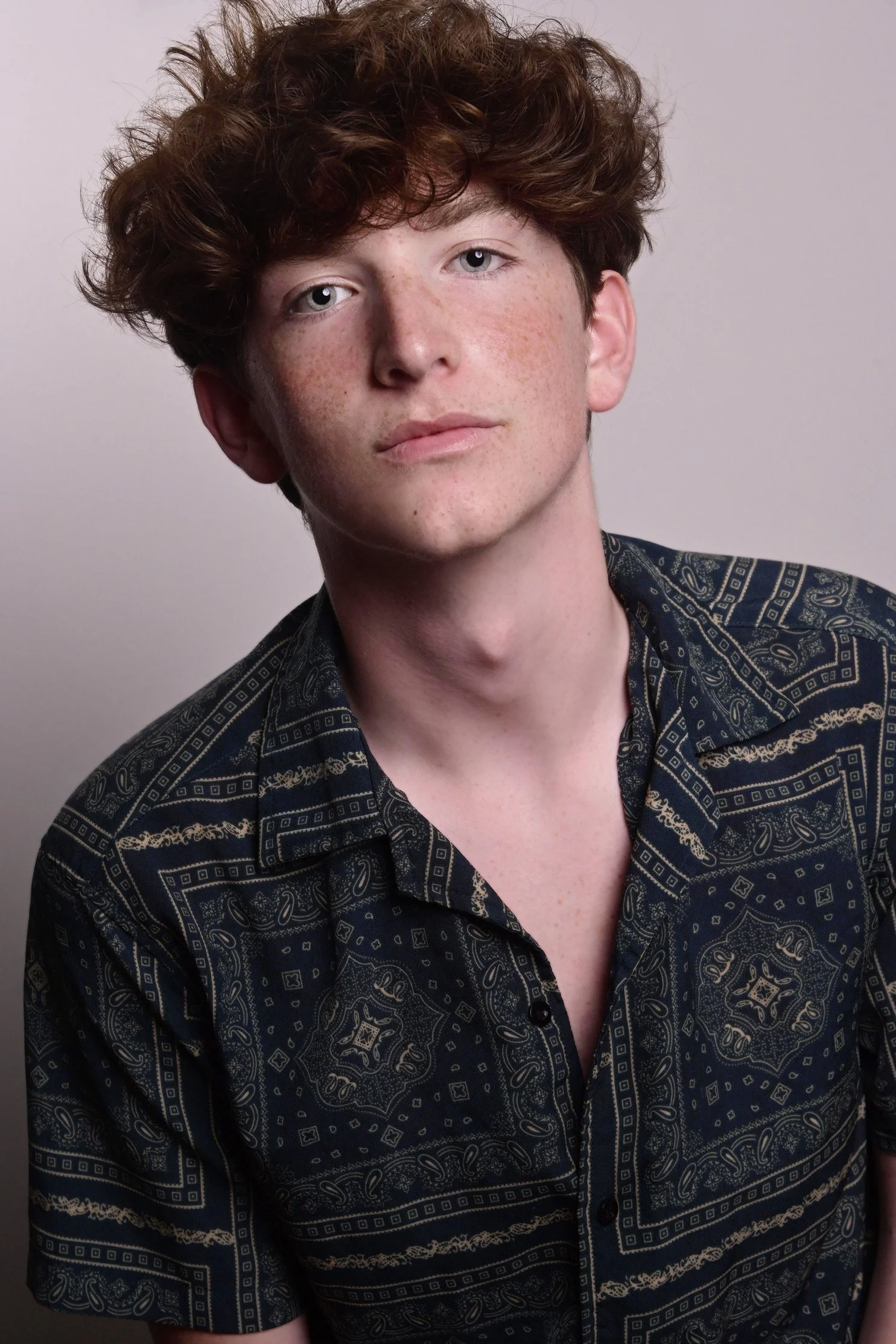 Portrait of a young man with wavy red hair, fair skin with freckles, wearing a dark patterned shirt against a light background.