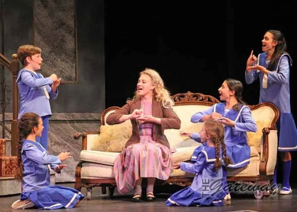 A woman dressed in vintage clothing sitting on a sofa with six young girls in matching blue costumes around her, engaging in a theatrical performance on a stage.