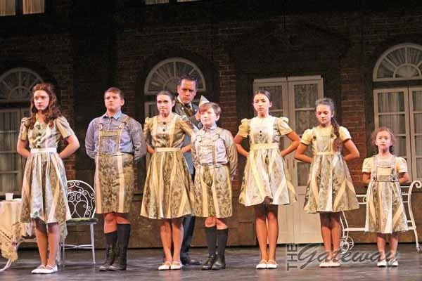 Seven girls and two boys in vintage dresses and shirts, standing in a line on stage with a brick building backdrop.