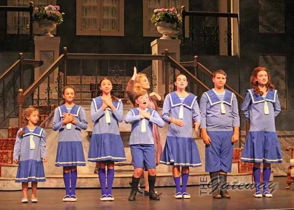 Children in blue sailor costumes performing on stage, with a woman directing them.
