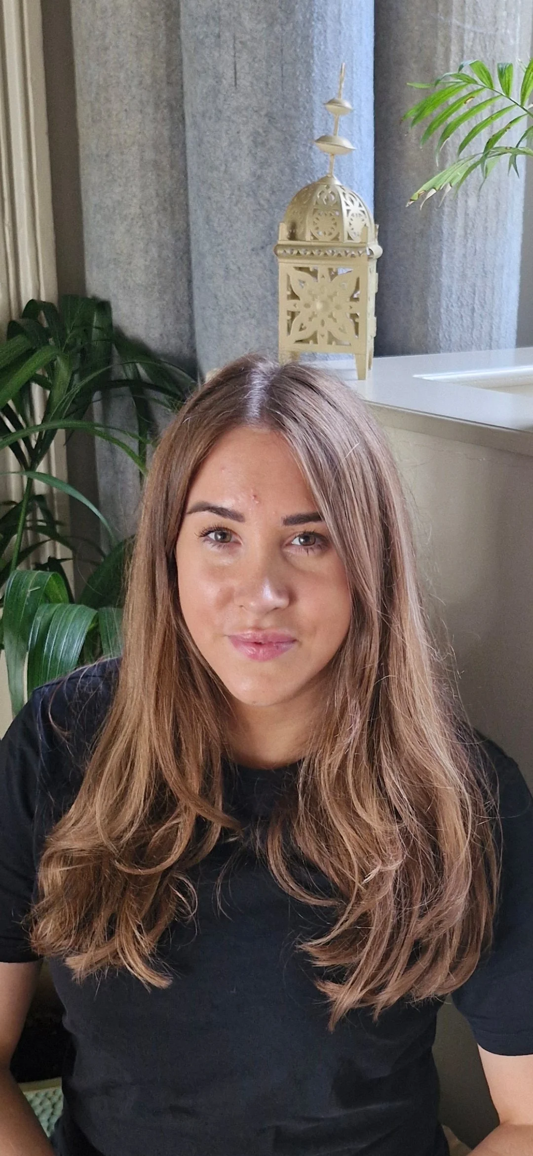 A young woman with long, wavy brown hair, wearing a black shirt, sitting indoors near a window with green plants and decorative objects in the background.