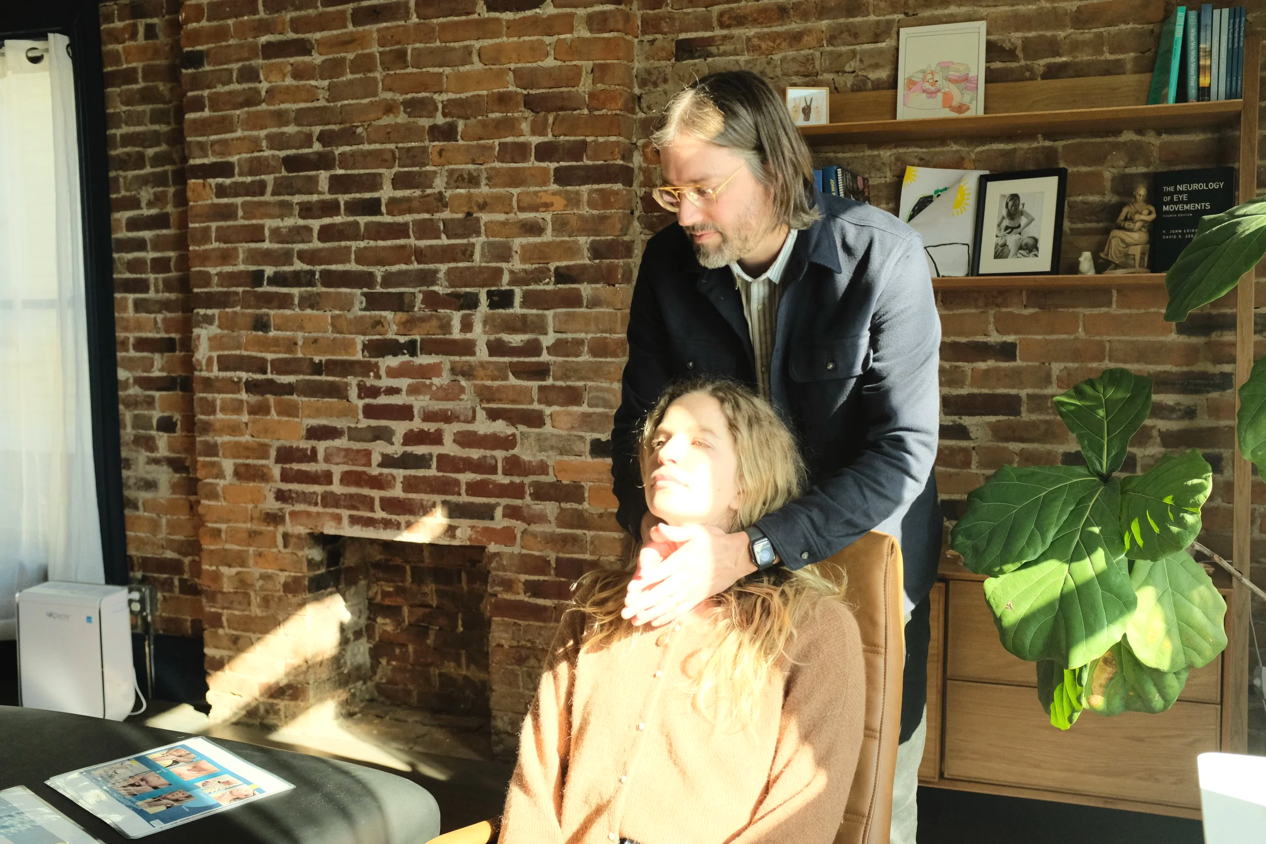 A man appears to be giving a neck massage or medical assessment to a young woman with long, curly hair in a cozy room with an exposed brick wall. Sunlight streams into the room, illuminating the woman’s face. There are books, framed photos, and artwork on the wooden shelf behind them, and a large leafy plant is visible on the right side of the image.