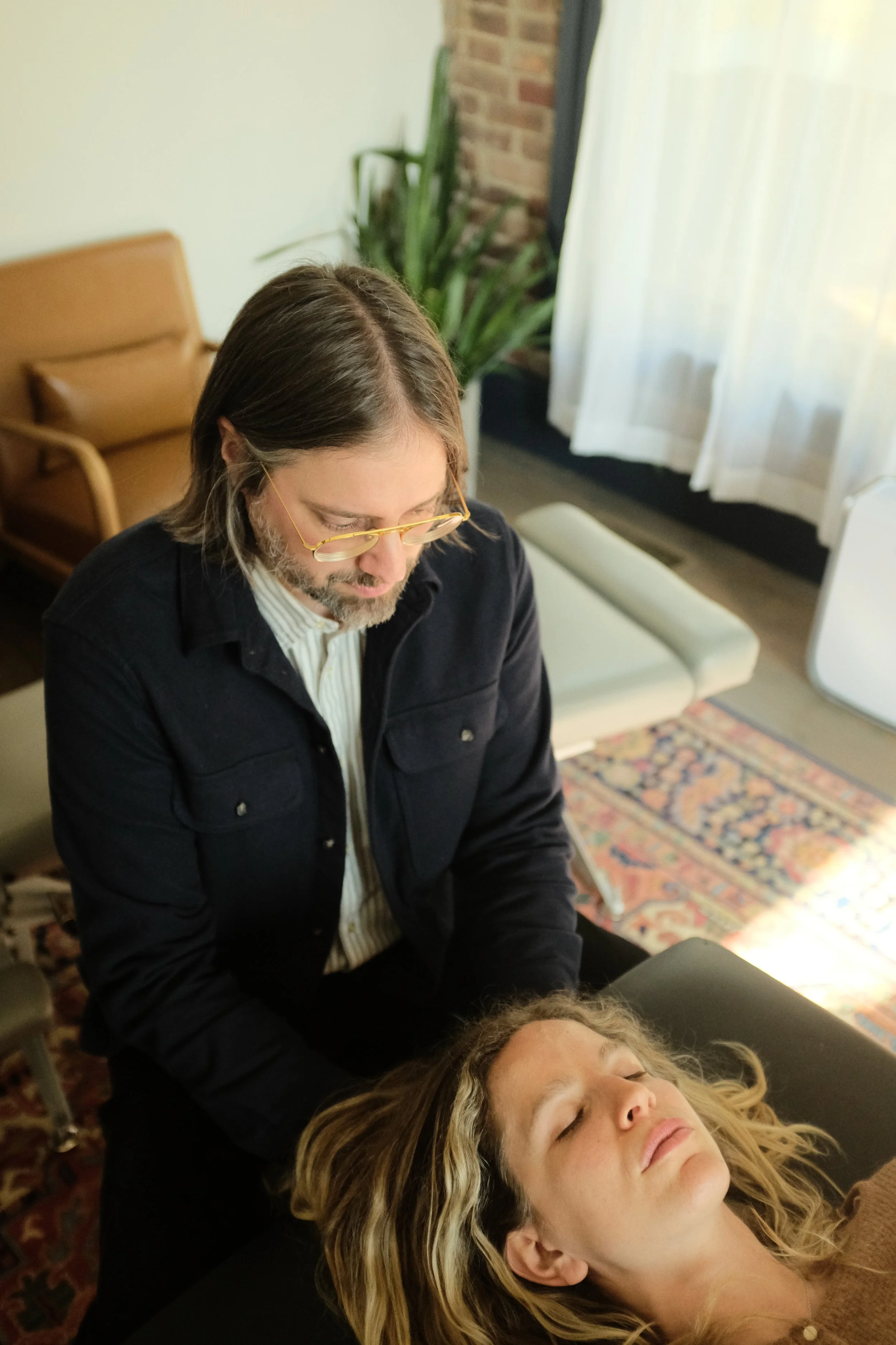 A man with glasses performs cranial therapy on a woman lying on a massage table in a cozy, well-lit room with a patterned rug and green plants.