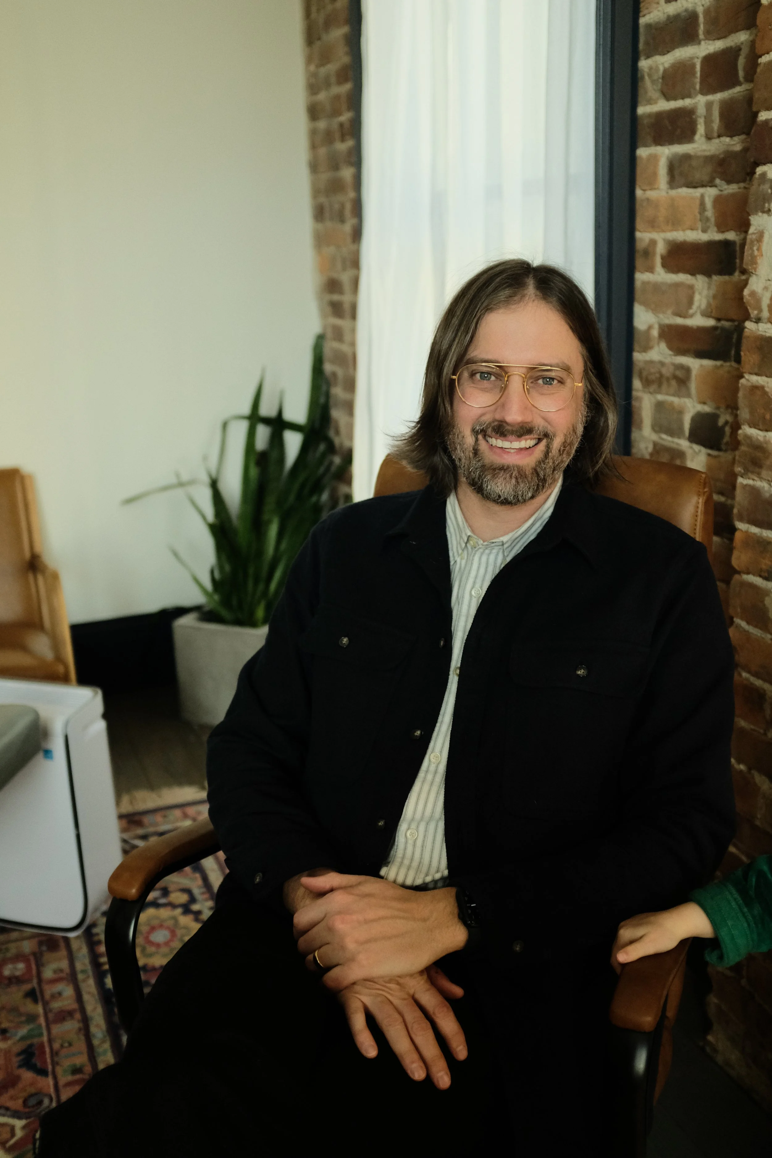 A man with shoulder-length hair, glasses, and a beard, sitting in a chair with a smile, against a background of a brick wall and a tall window with white curtains.