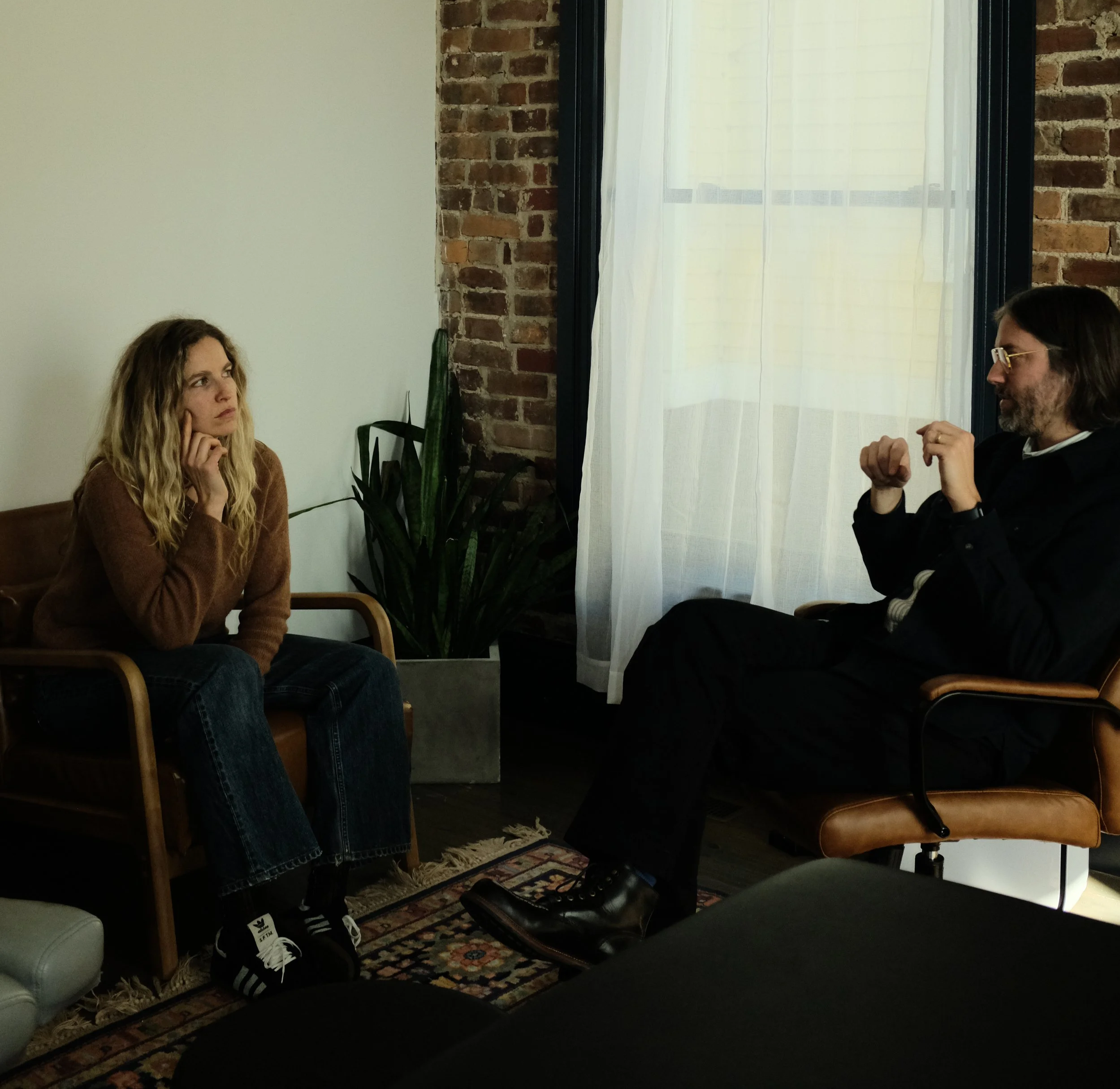 A woman sits in a chair, listening carefully, while a man talks with gestures in a room with exposed brick and a large window with white curtains.