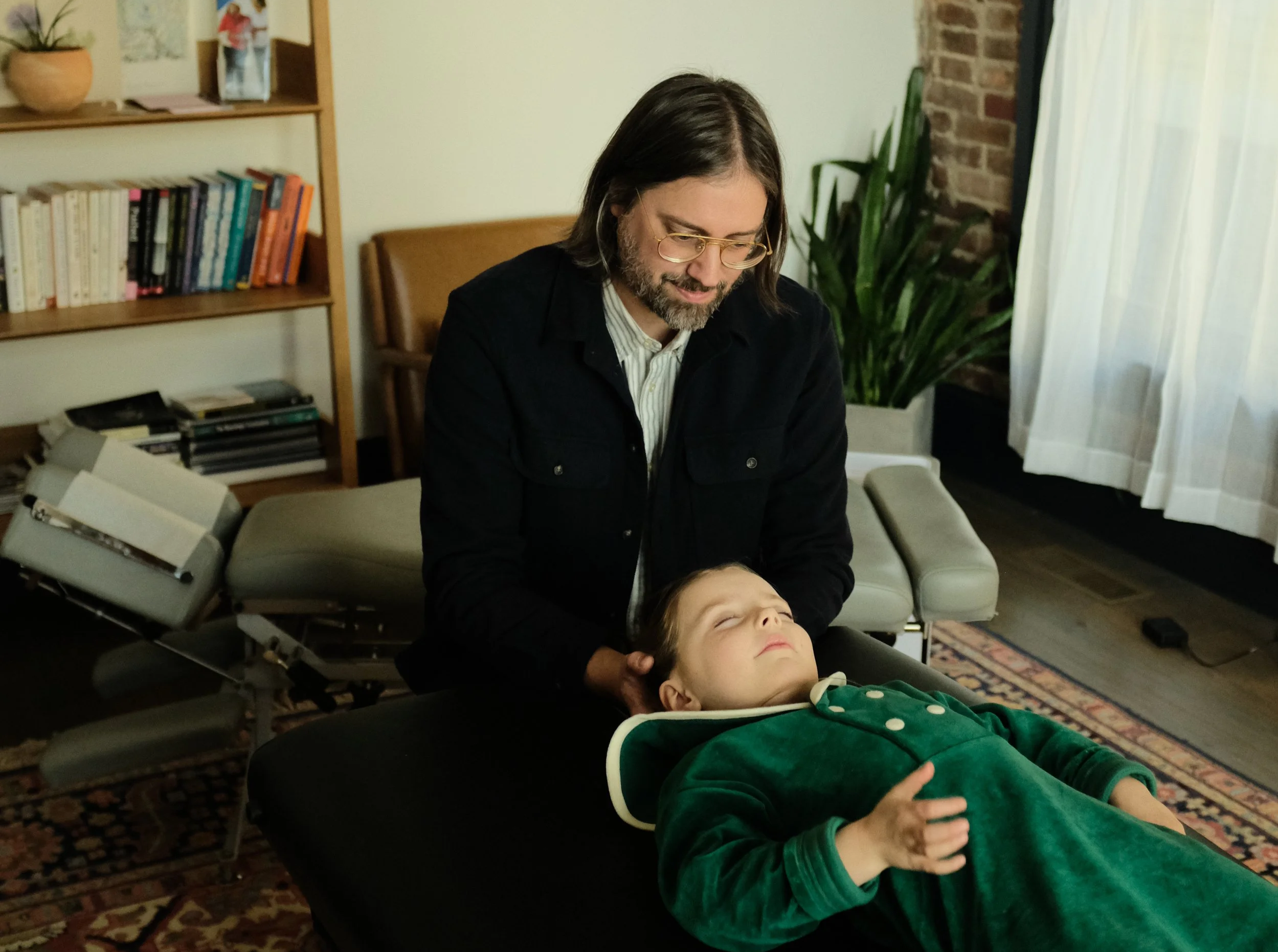 A man with shoulder-length hair, glasses, and a beard provides a chiropractic adjustment to a young girl lying on a chiropractic table. The scene takes place in a cozy, well-lit room with a bookshelf, a large plant, and a window with curtains.