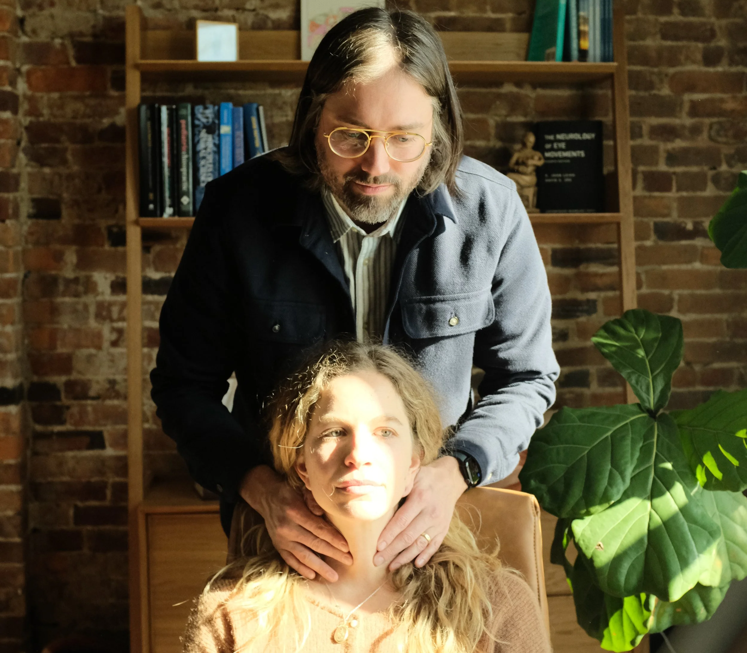 A man with long hair and glasses giving a neck massage to a woman with blonde hair sitting in a chair. They are indoors against a brick wall with a bookshelf and a large green plant nearby.