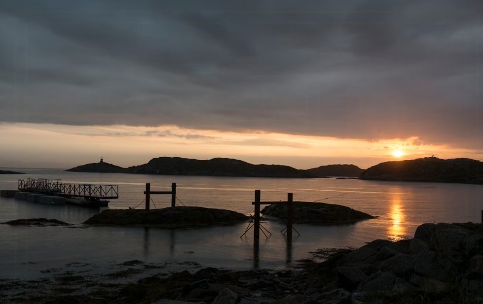 Sunset over a calm body of water with rocky islands, a lighthouse in the distance, and a dock on the left.