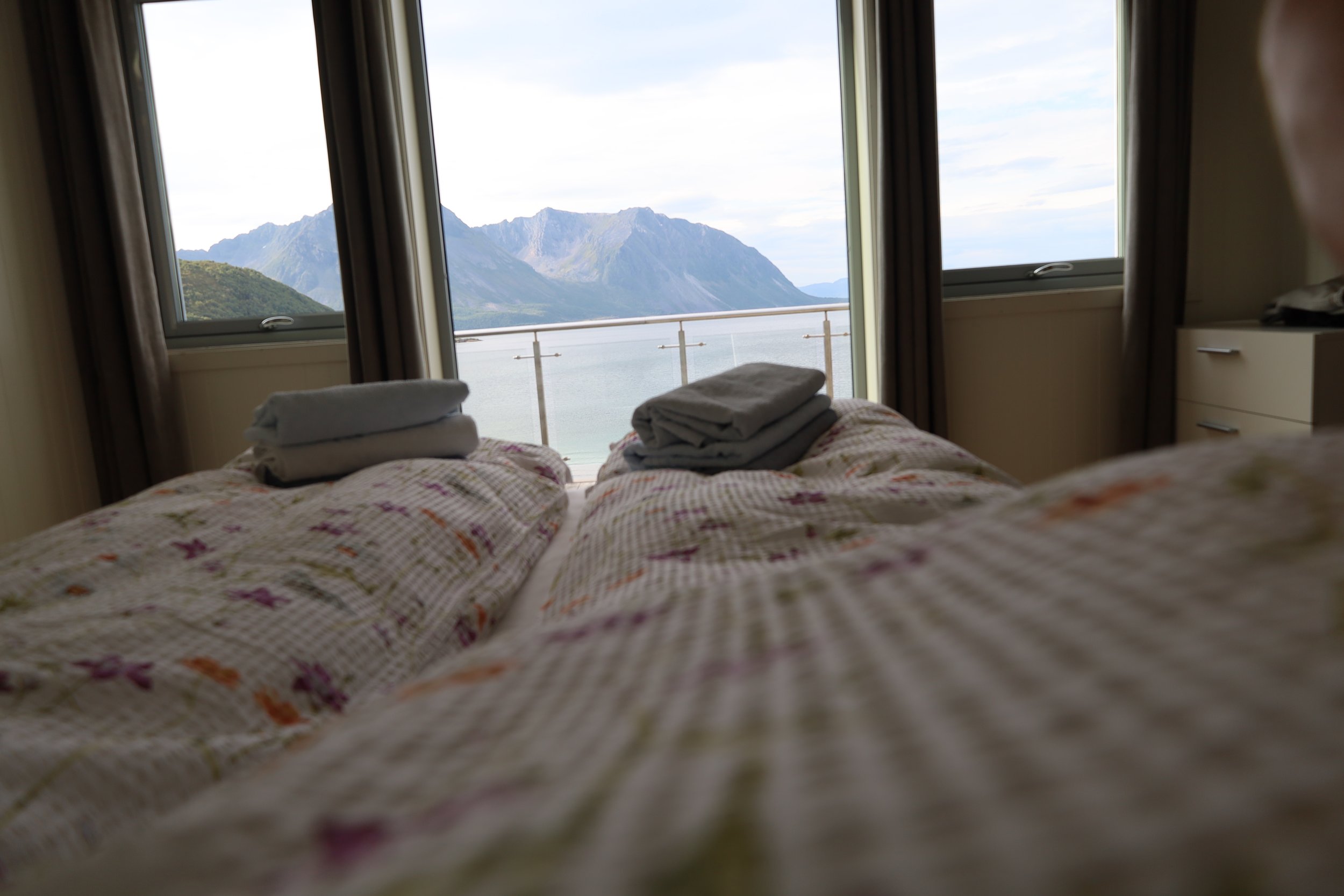 A cozy bedroom with a view of mountains and water through large windows, with folded towels on the bed.
