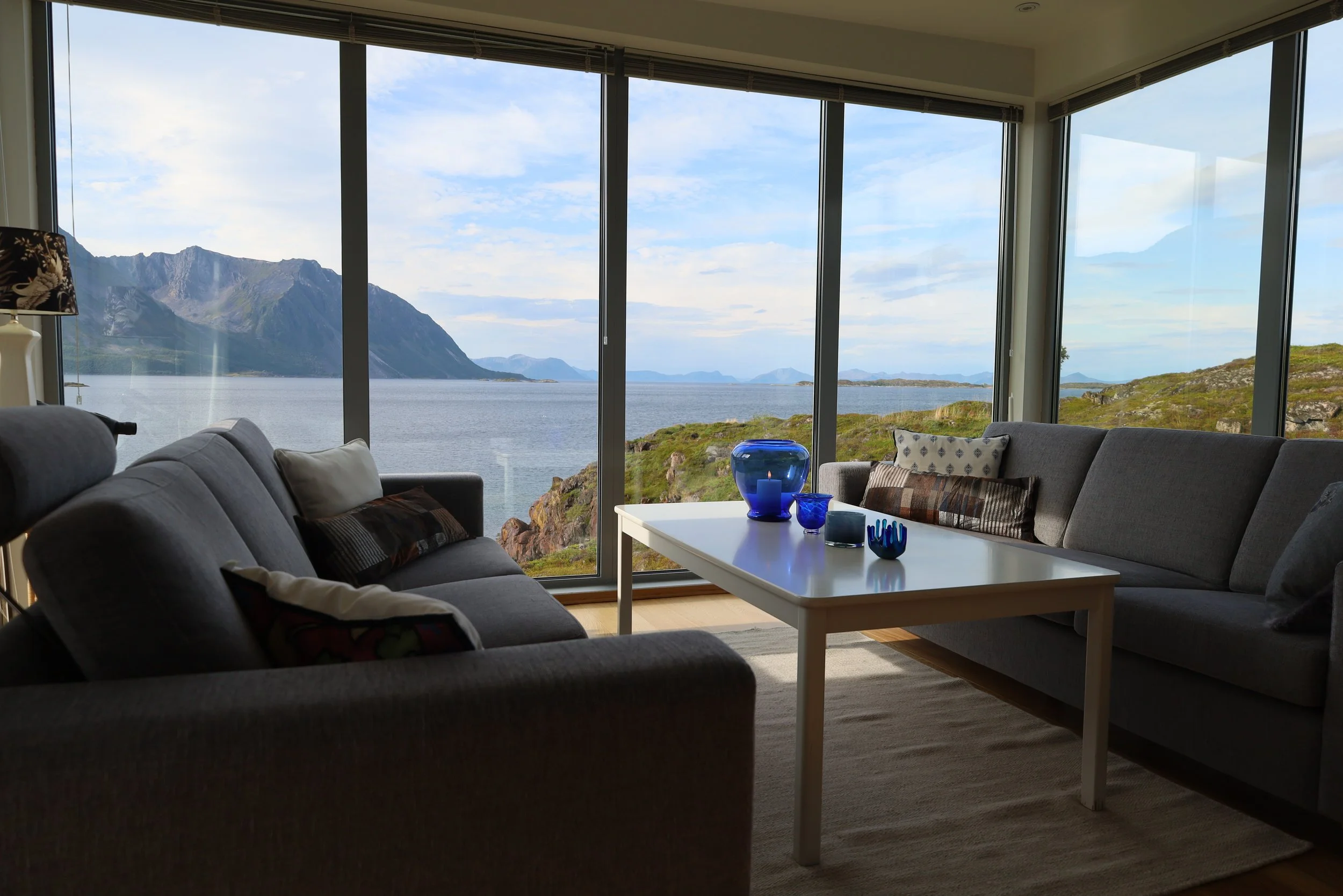 Living room with large windows showing mountains, water, and sky outside, furnished with gray sofas, a white coffee table with blue glass decor, and a beige rug.
