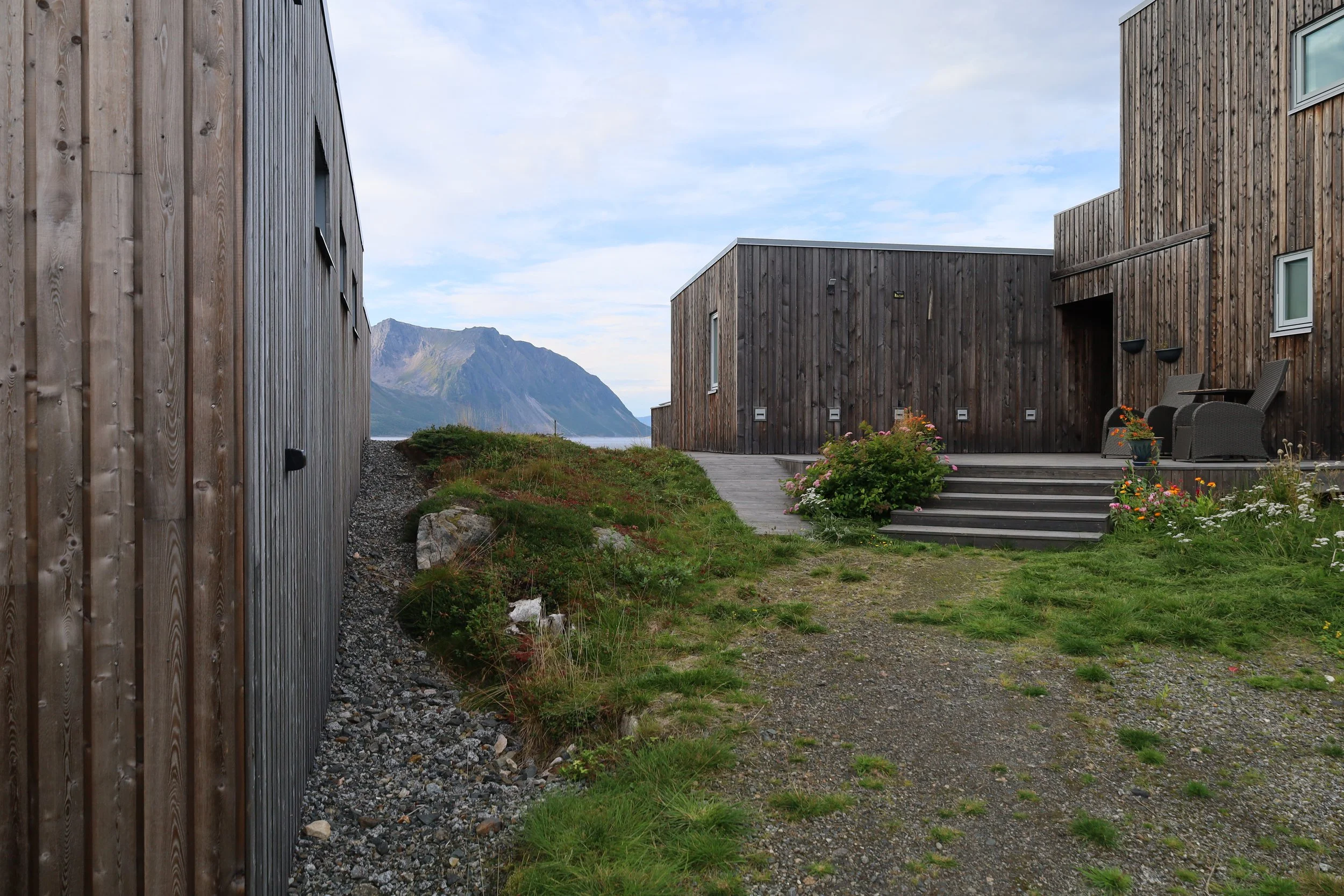 Two modern wooden houses with a terrace, outdoor chairs, and plants, set against a mountain landscape with a partly cloudy sky.