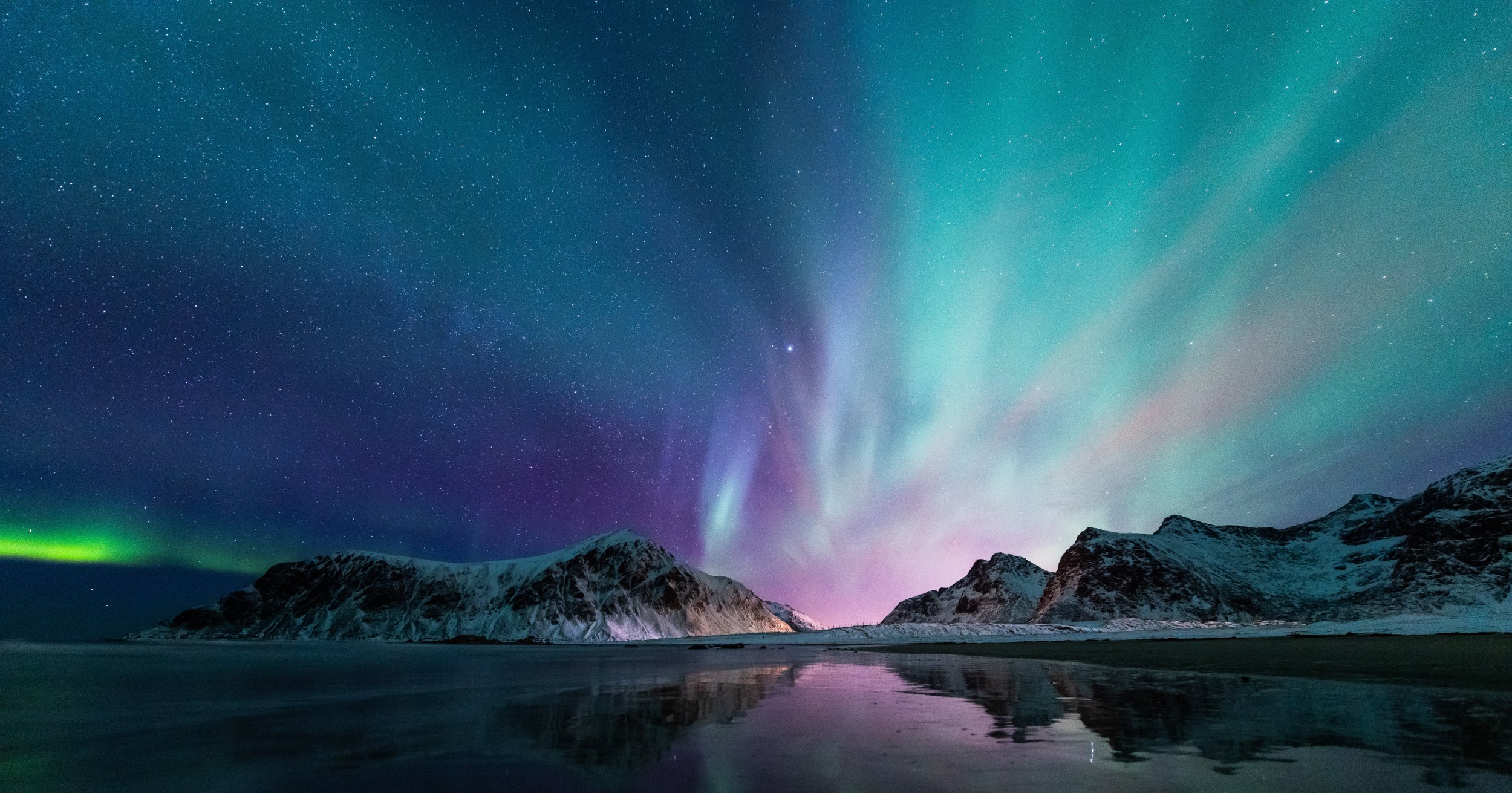 Nighttime landscape showing snow-covered mountains along the shoreline, with a colorful aurora borealis and stars in the sky above.