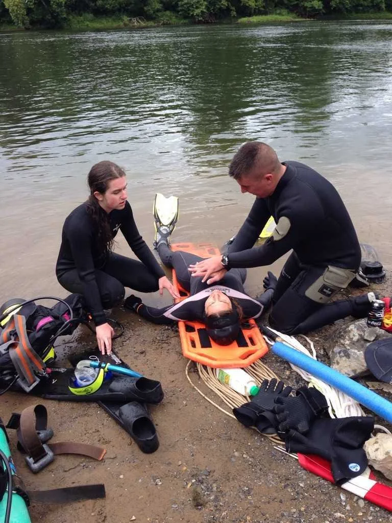 Rescue team assists a person lying on a stretcher by a riverbank, surrounded by rescue equipment and gear.