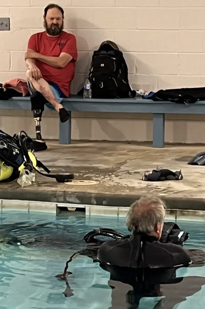 A man with a prosthetic leg sitting on a bench at an indoor swimming pool, next to a backpack and water bottles, with a woman in the water wearing a scuba diving suit nearby.