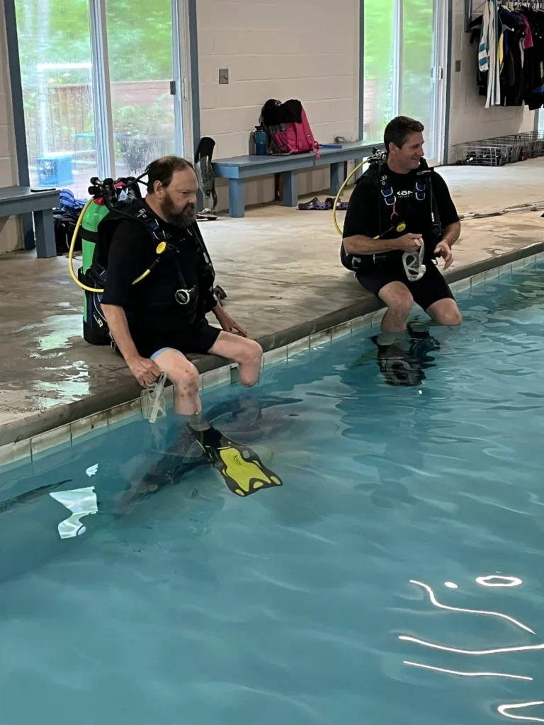 Two men sitting on the edge of an indoor swimming pool wearing scuba gear, with their feet in the water.
