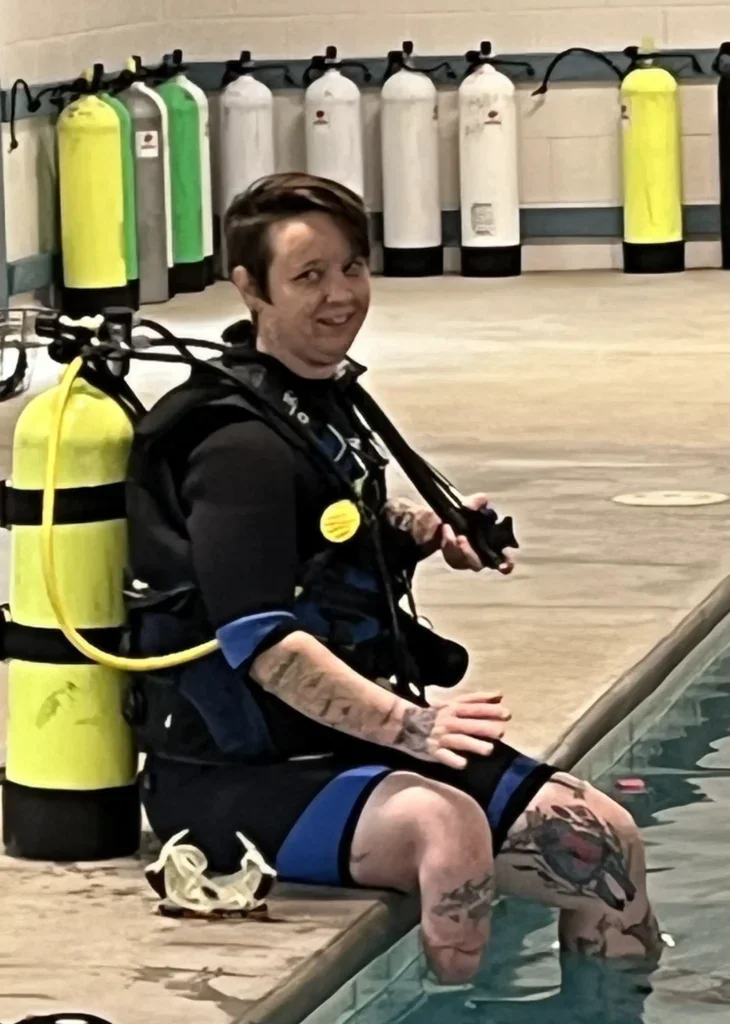 A woman in scuba gear sitting on the edge of a swimming pool with her feet in the water, smiling at the camera, with oxygen tanks lined up in the background.