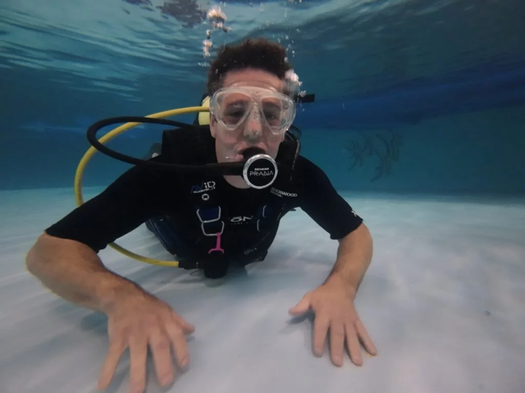 Person scuba diving underwater with scuba gear, arms extended, and a blurry fish in the background.