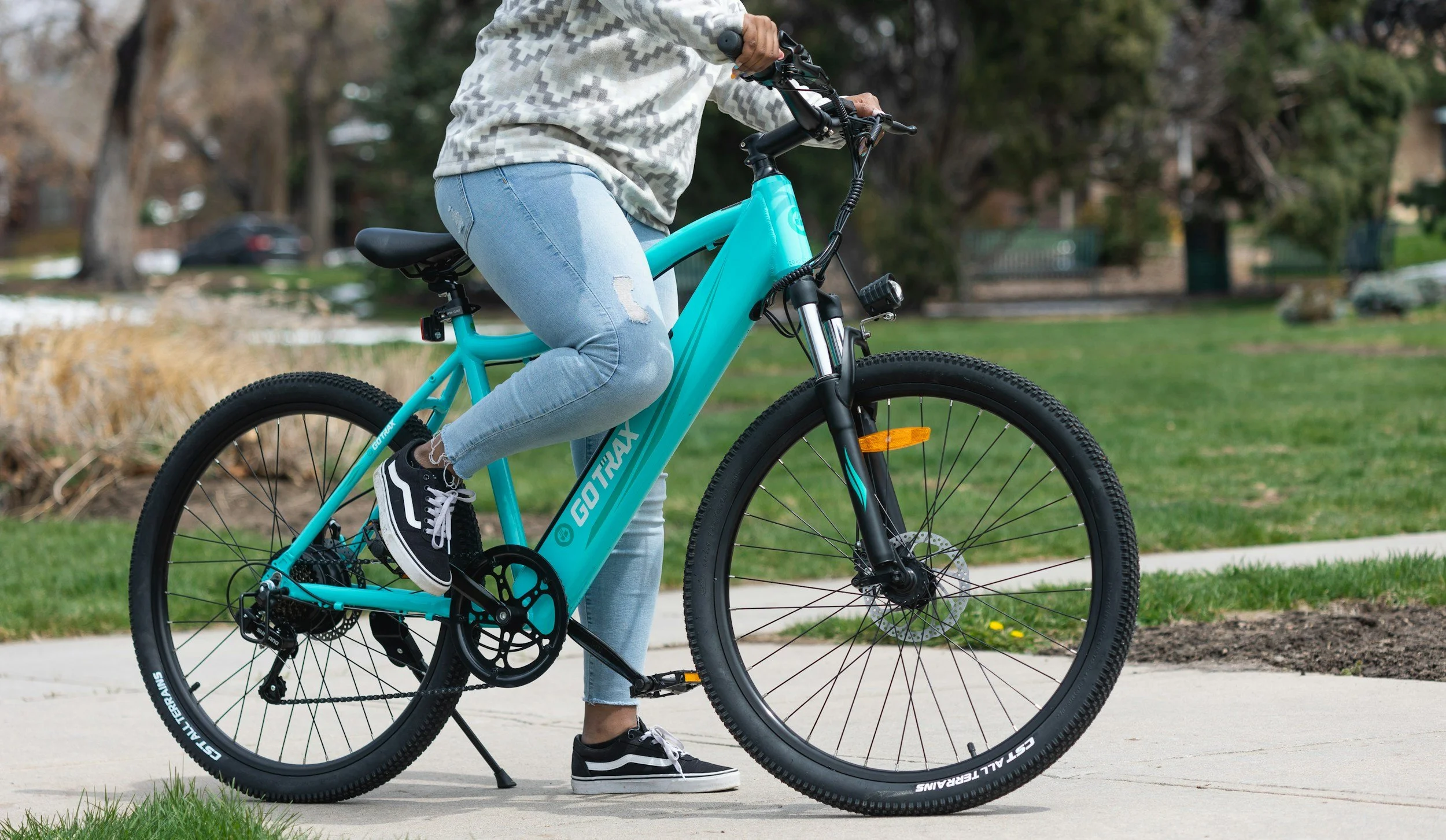 Person riding a turquoise Gotype electric bicycle on a sidewalk in a park, wearing jeans and sneakers.