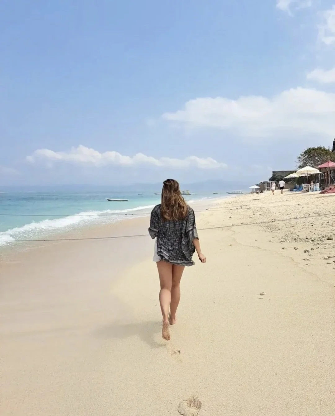 A woman walking barefoot on a sandy beach with clear blue water and umbrellas in the background.