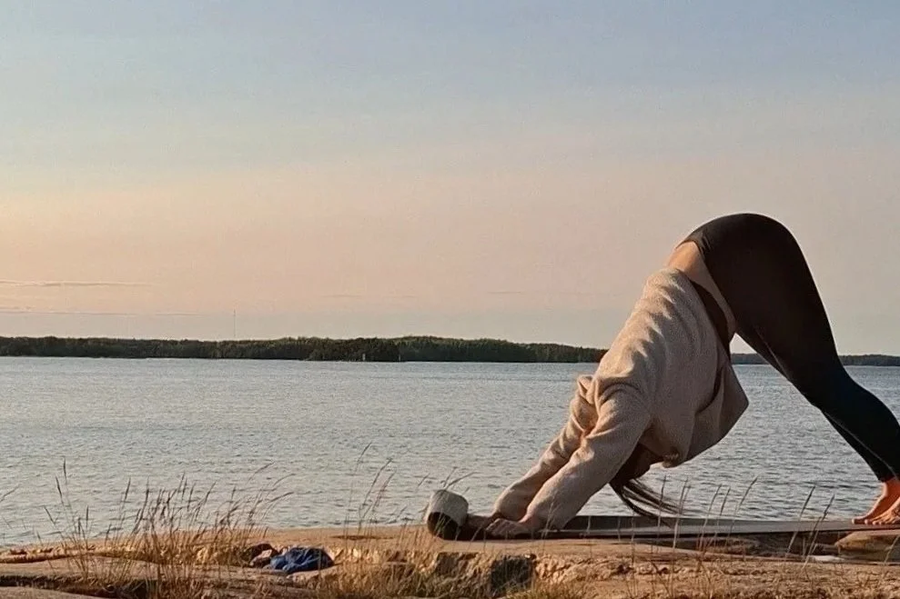 Person practicing yoga outdoors on a beach near water during sunset, in a downward dog pose.
