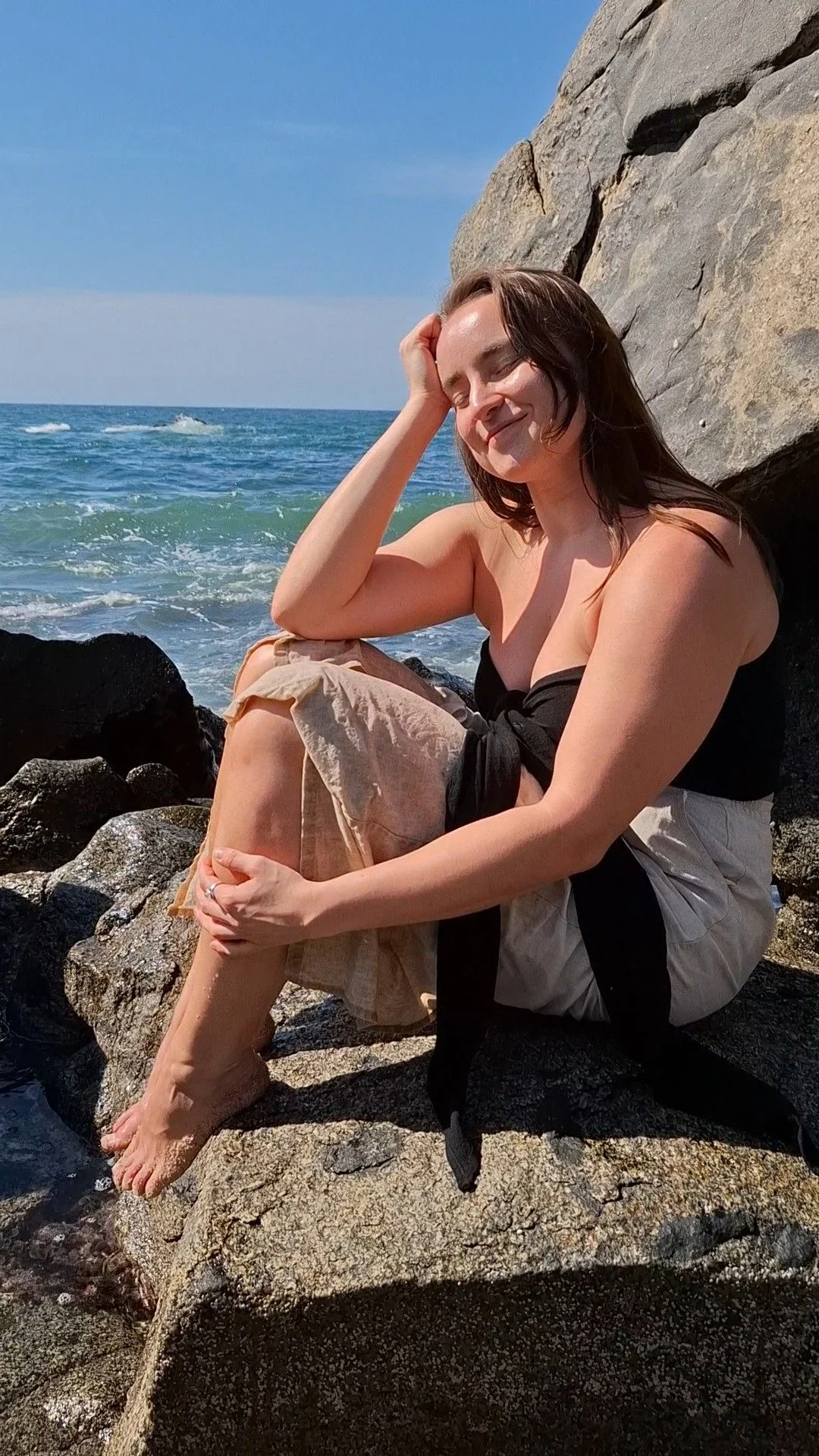 A woman sitting on rocks by the ocean, smiling with eyes closed, wearing a black strapless top and beige pants, with her feet in the water and a large rock behind her.