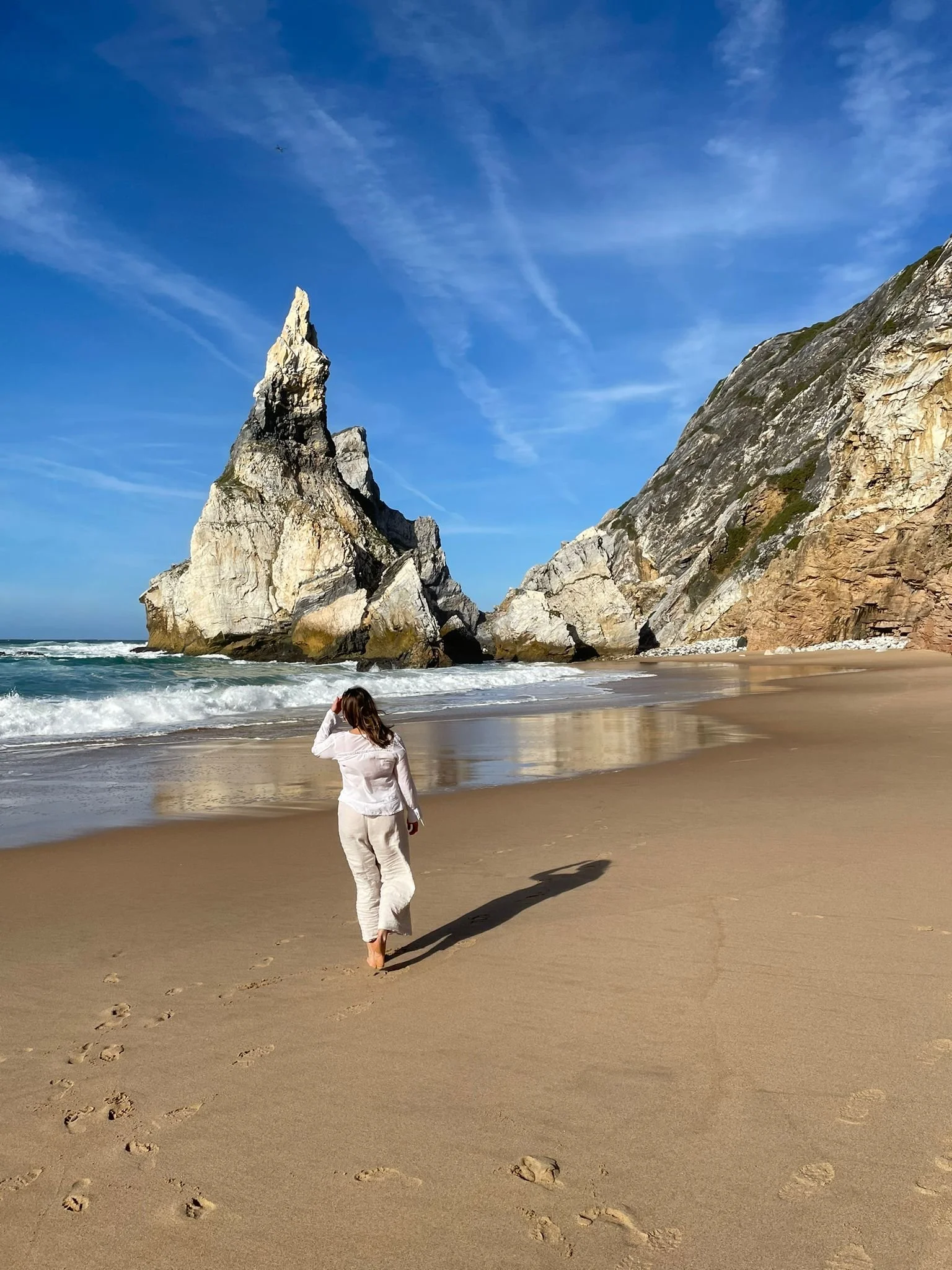 A woman walking barefoot on a sandy beach with footprints, towards large rocks and cliffs, under a blue sky with wispy clouds.