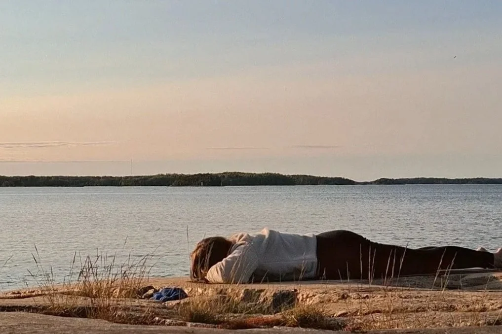 Person lying on a sandy beach near a body of water at sunset, with a small patch of grass and rocks nearby.
