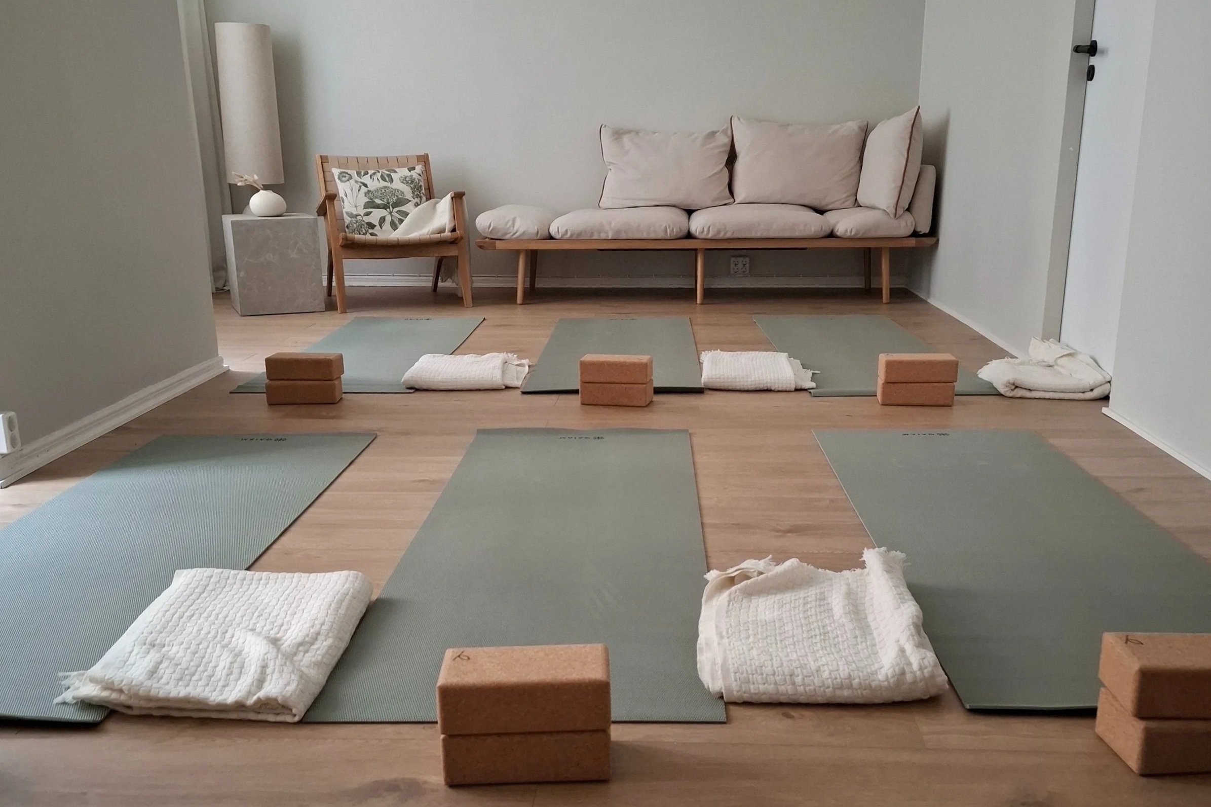 Yoga mats, blocks, and blankets set up in a spacious room with a sitting area featuring a beige sofa and a wooden chair with a pillow.