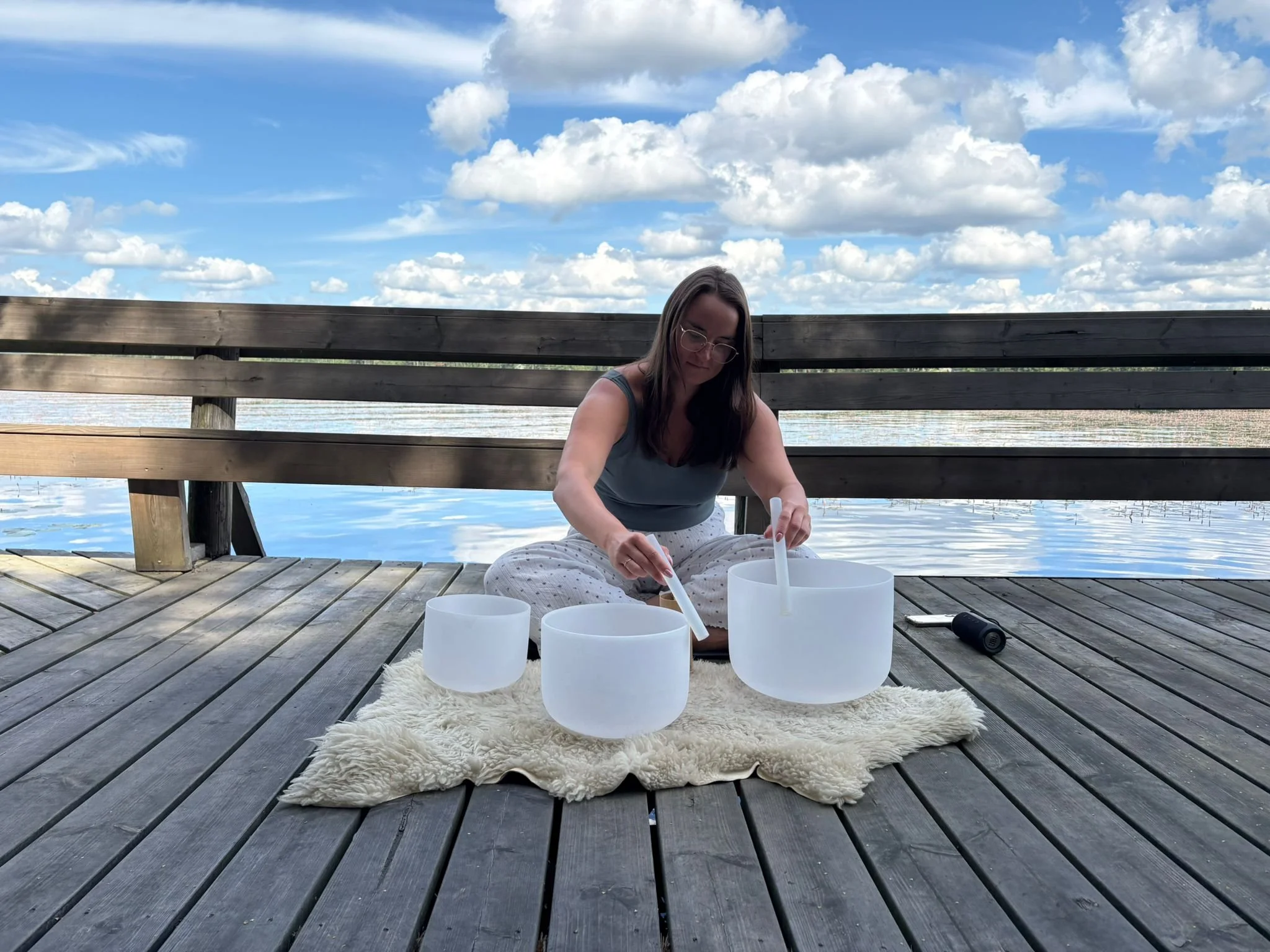 A woman sitting on a wooden dock by the water playing crystal singing bowls during daytime with a partly cloudy sky.