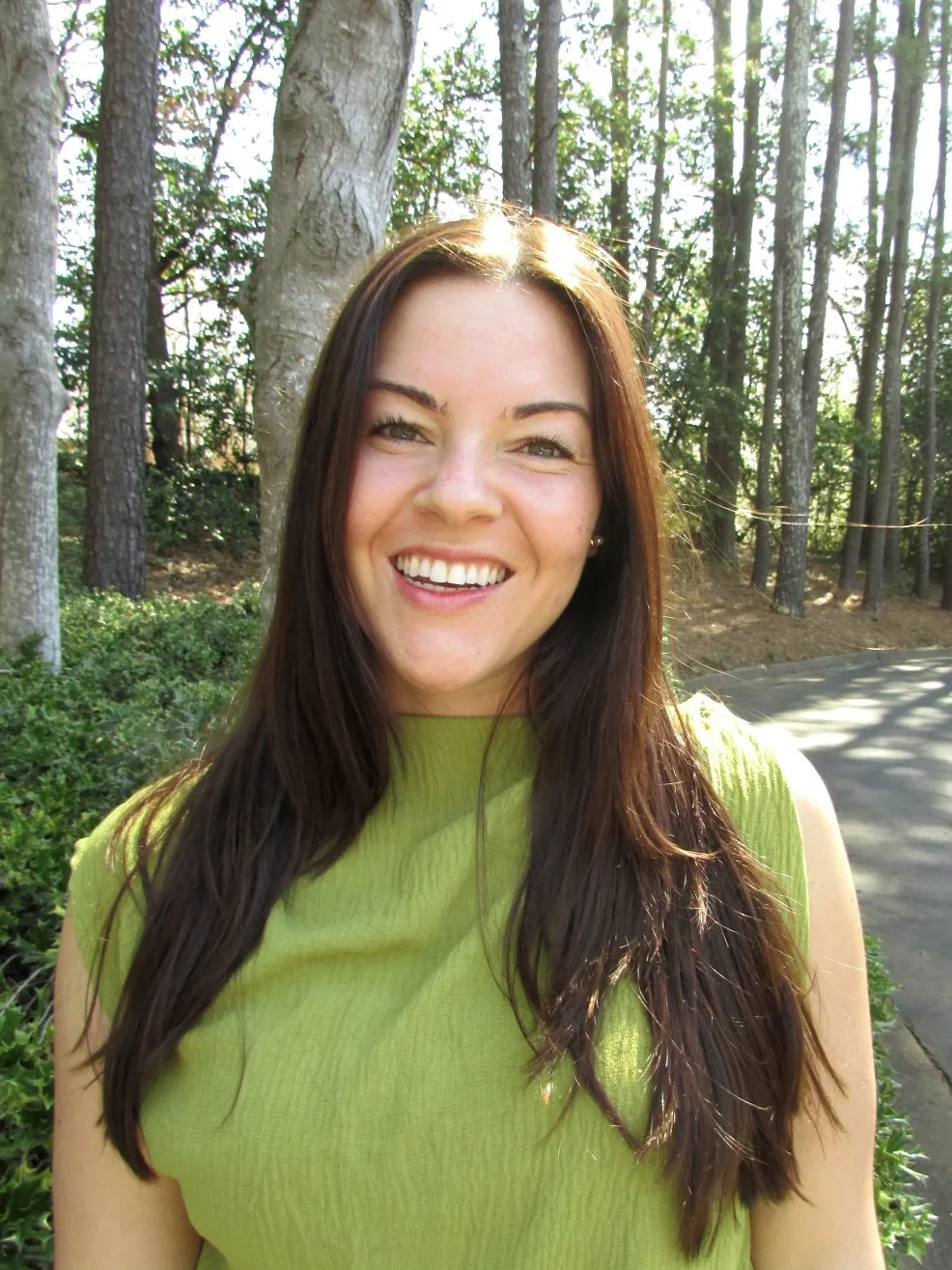 Young woman with long brown hair smiling outdoors in a green sleeveless top, standing in front of trees and greenery.