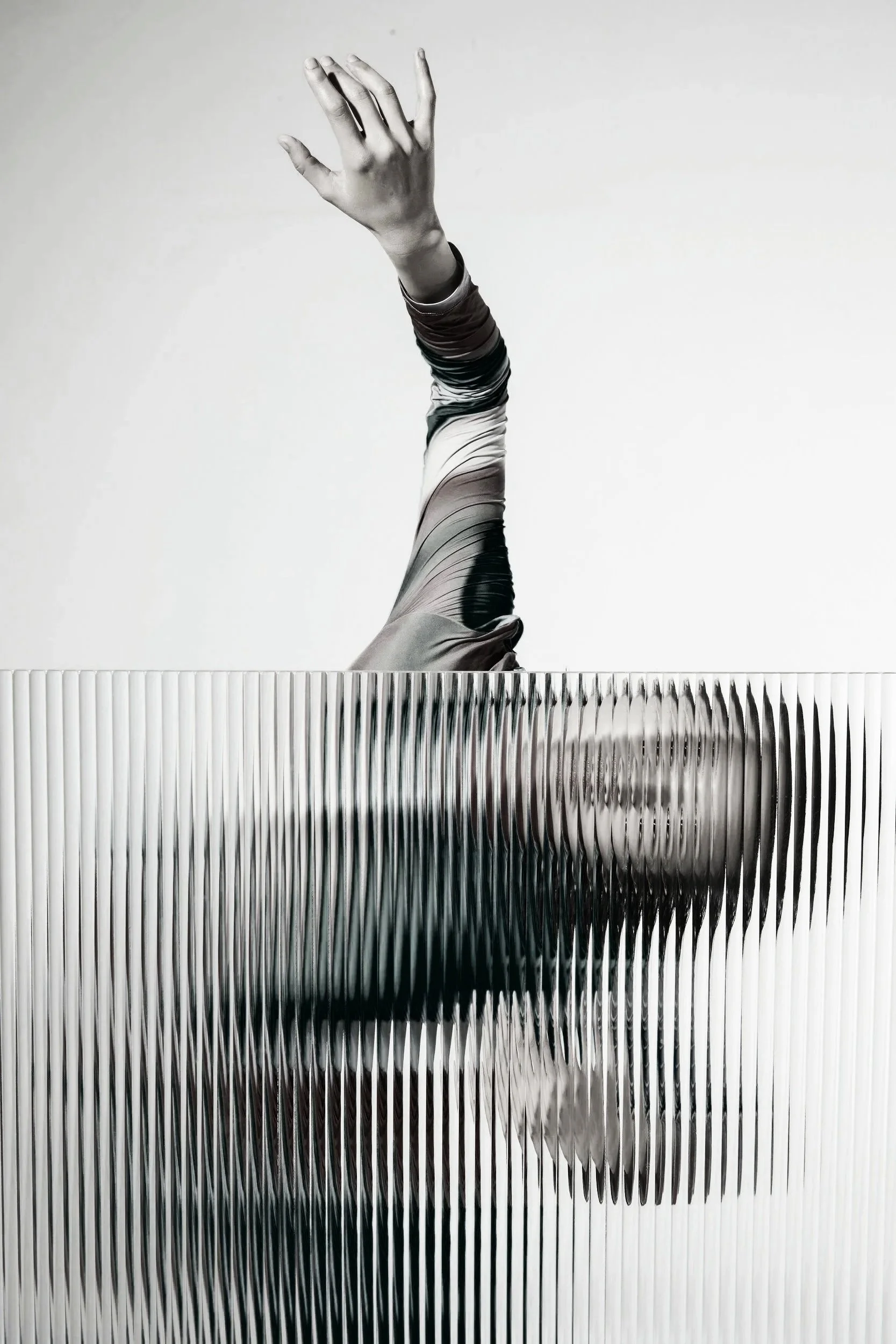 A black and white photograph of a woman with her head and upper body obscured behind a textured glass panel, with her arm extended upward and her hand reaching out.