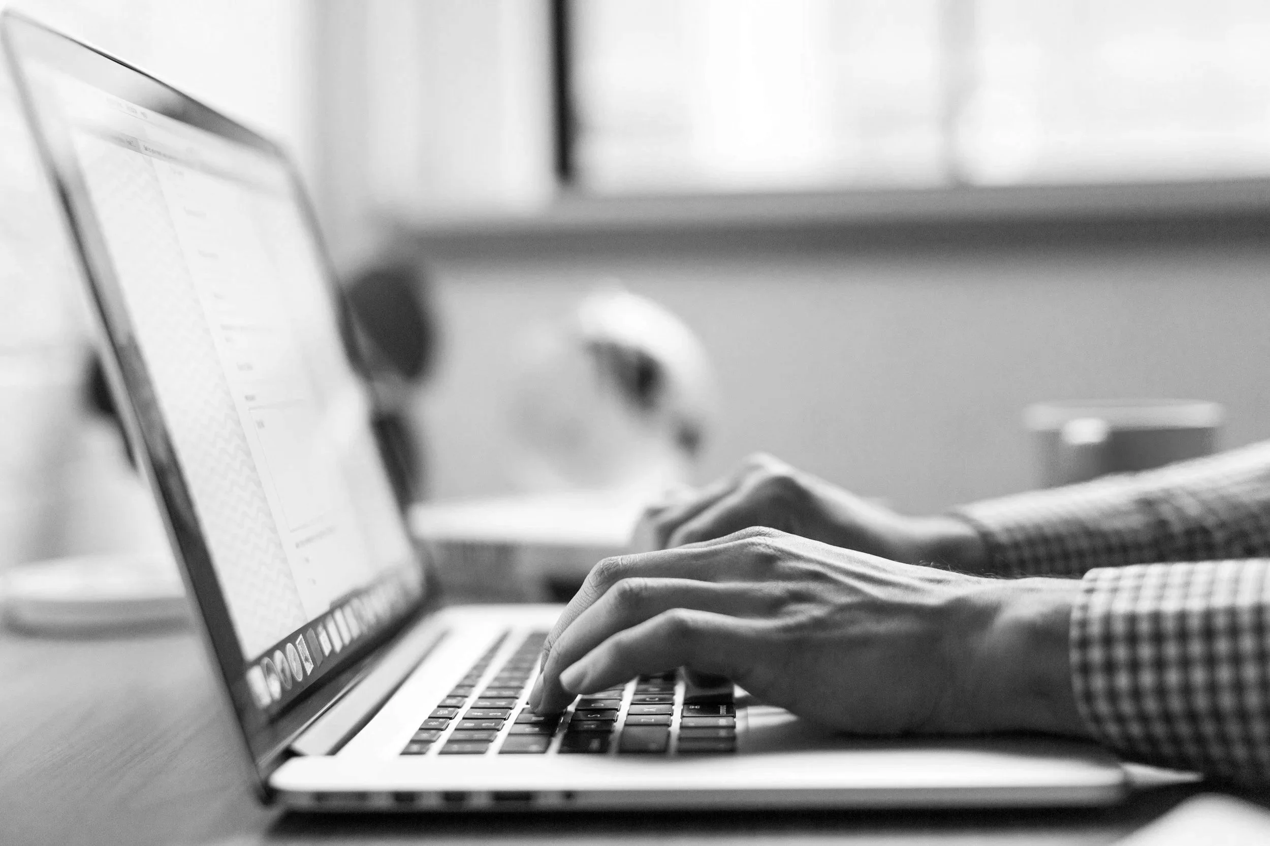 Black and white photo of a person typing on a laptop keyboard, with a blurred background of a window and a mug.
