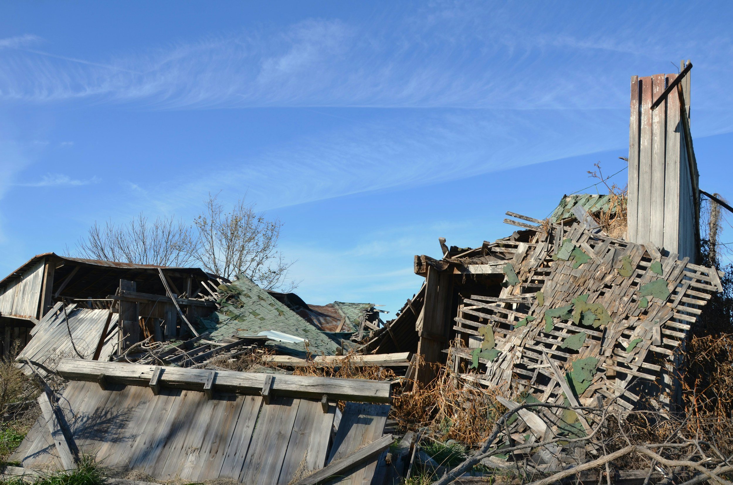 Ruined wooden building with collapsed structures and debris under a blue sky with wispy clouds.