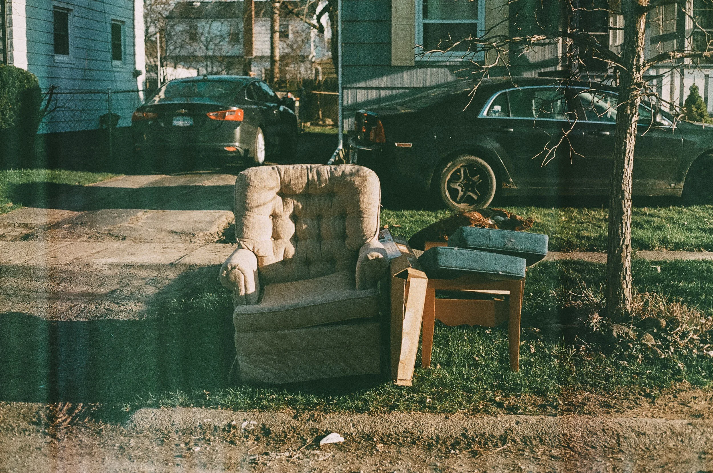 Old armchair and small side table with two boxes on top, outside in front of house, with two cars parked on the street, and a bare tree.