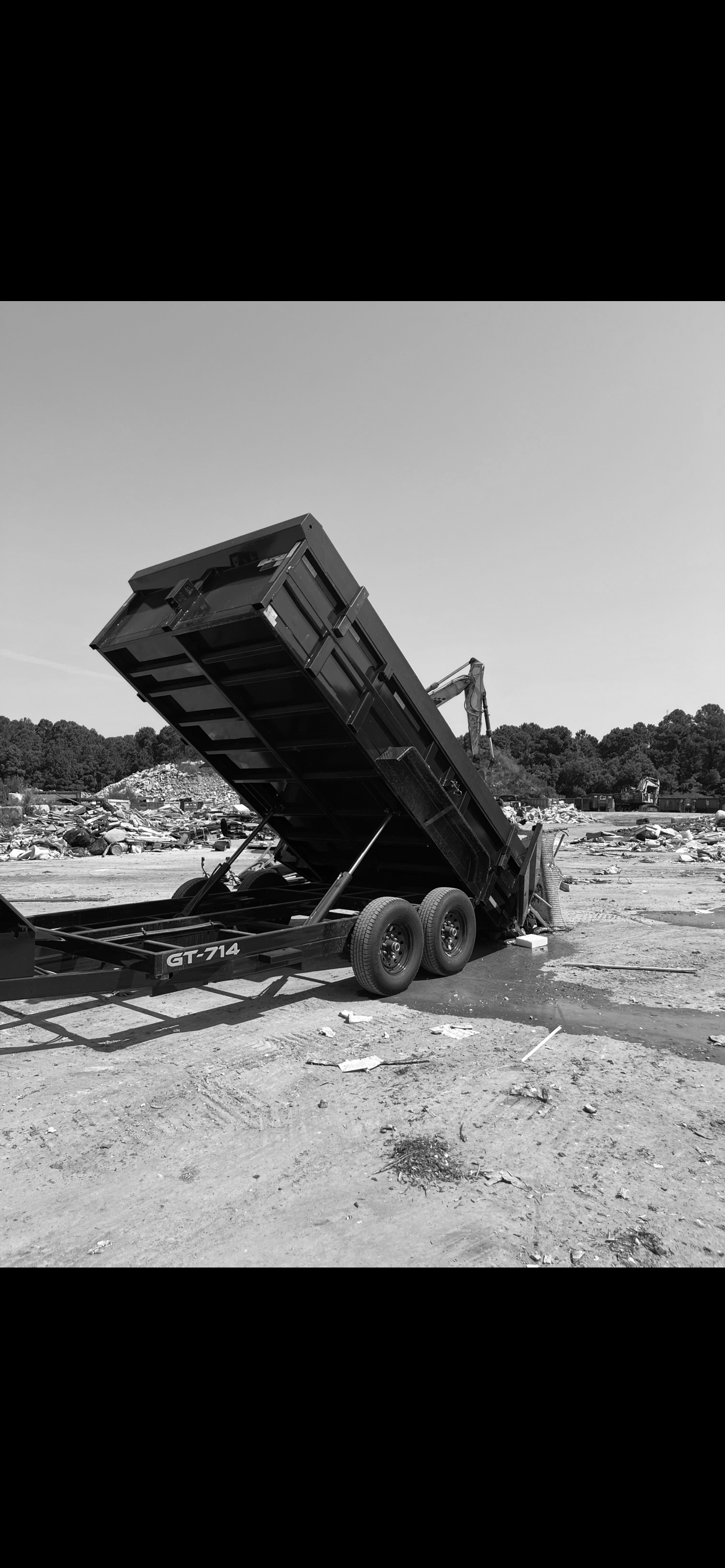 A black and white photo of a dump trailer in a scrapyard, with its bed raised.
