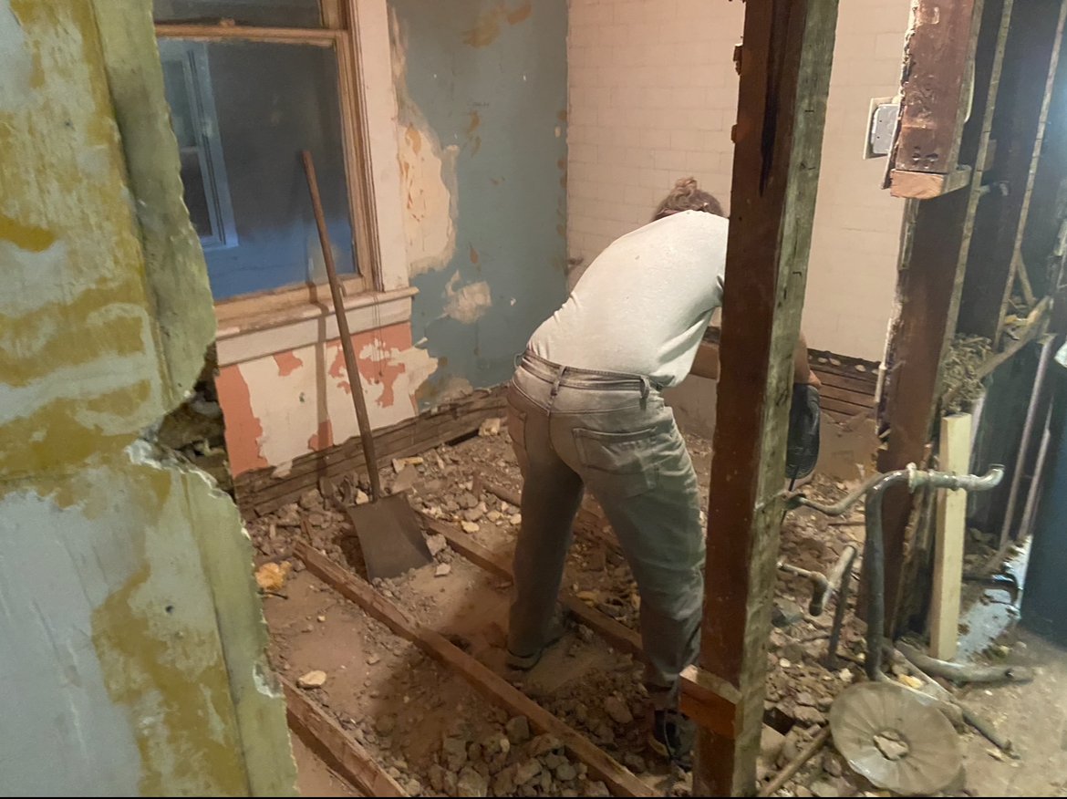 A person working on a construction or renovation project inside a building, lifting a wooden beam in a partially demolished or under-construction room with exposed walls and pipes.