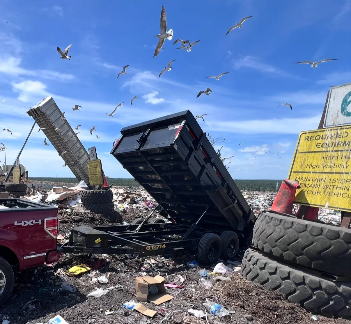 Debris filled landscape with overturned trailers, a red pickup truck, large tires, and birds flying in the sky under a partly cloudy blue sky.