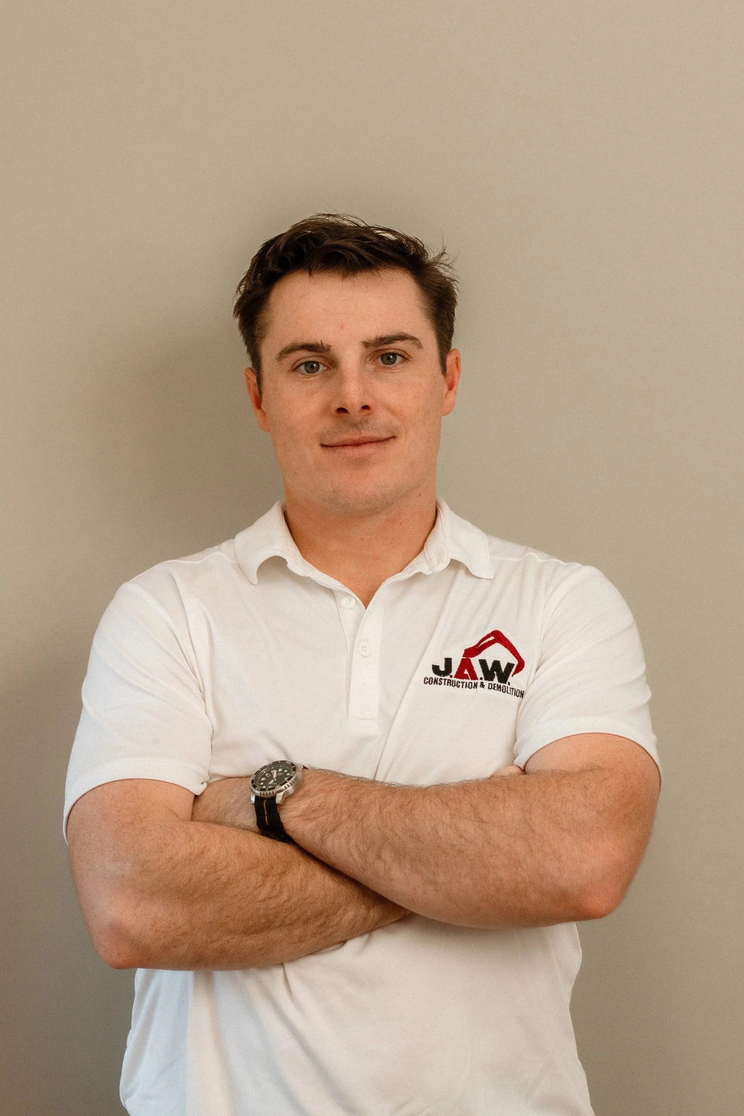 A young man with short brown hair and fair skin, standing against a plain beige wall, dressed in a white polo shirt with a logo that reads "JAW Construction & Demolition," with arms crossed and wearing a wristwatch.