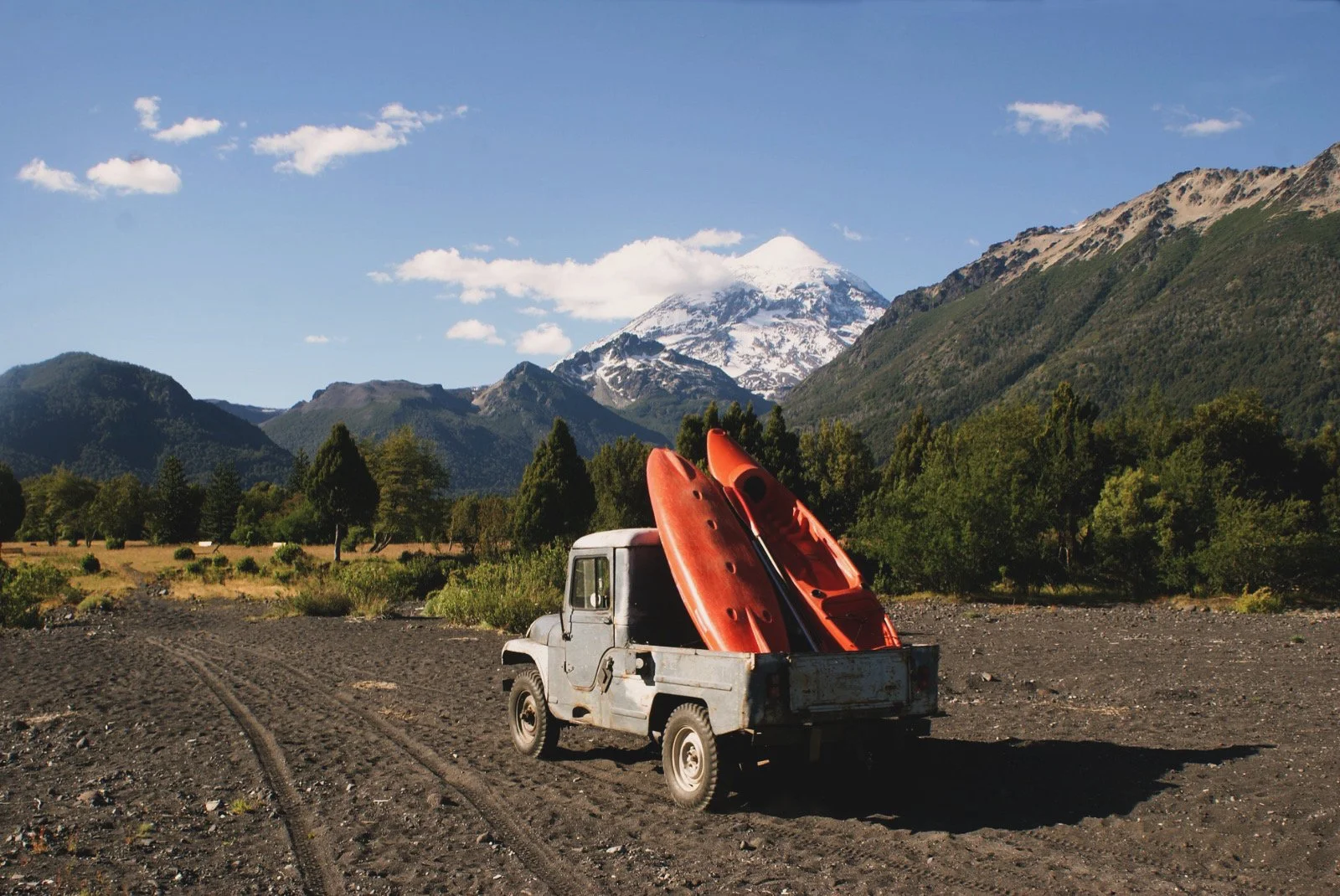 Camioneta antigua en un campo con dos kayaks anaranjados en su caja, montañas y volcán con nieve en el fondo bajo un cielo despejado.