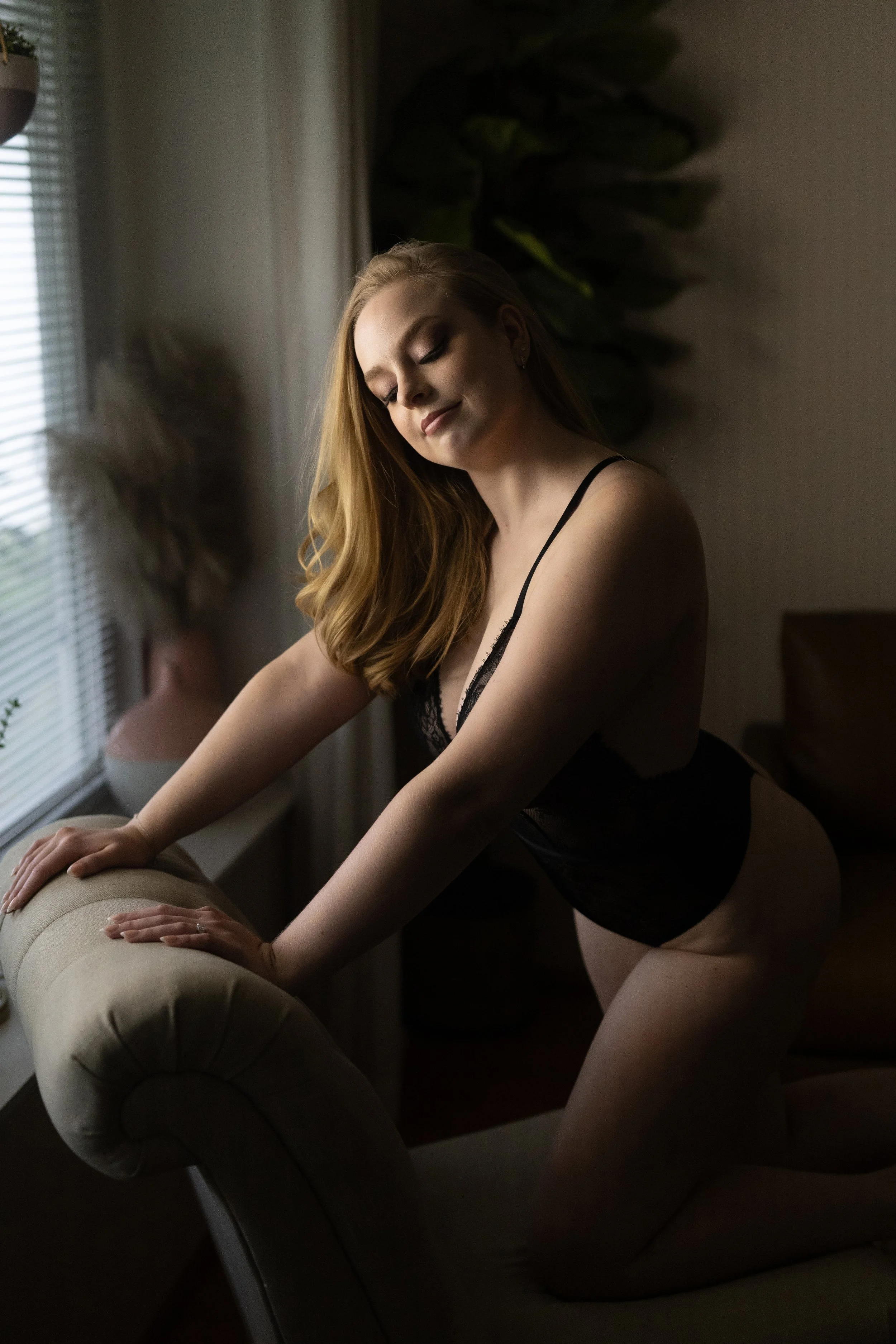 A woman with long red hair in black lingerie leaning over an upholstered armchair in a dimly lit room, with a window and plants in the background.