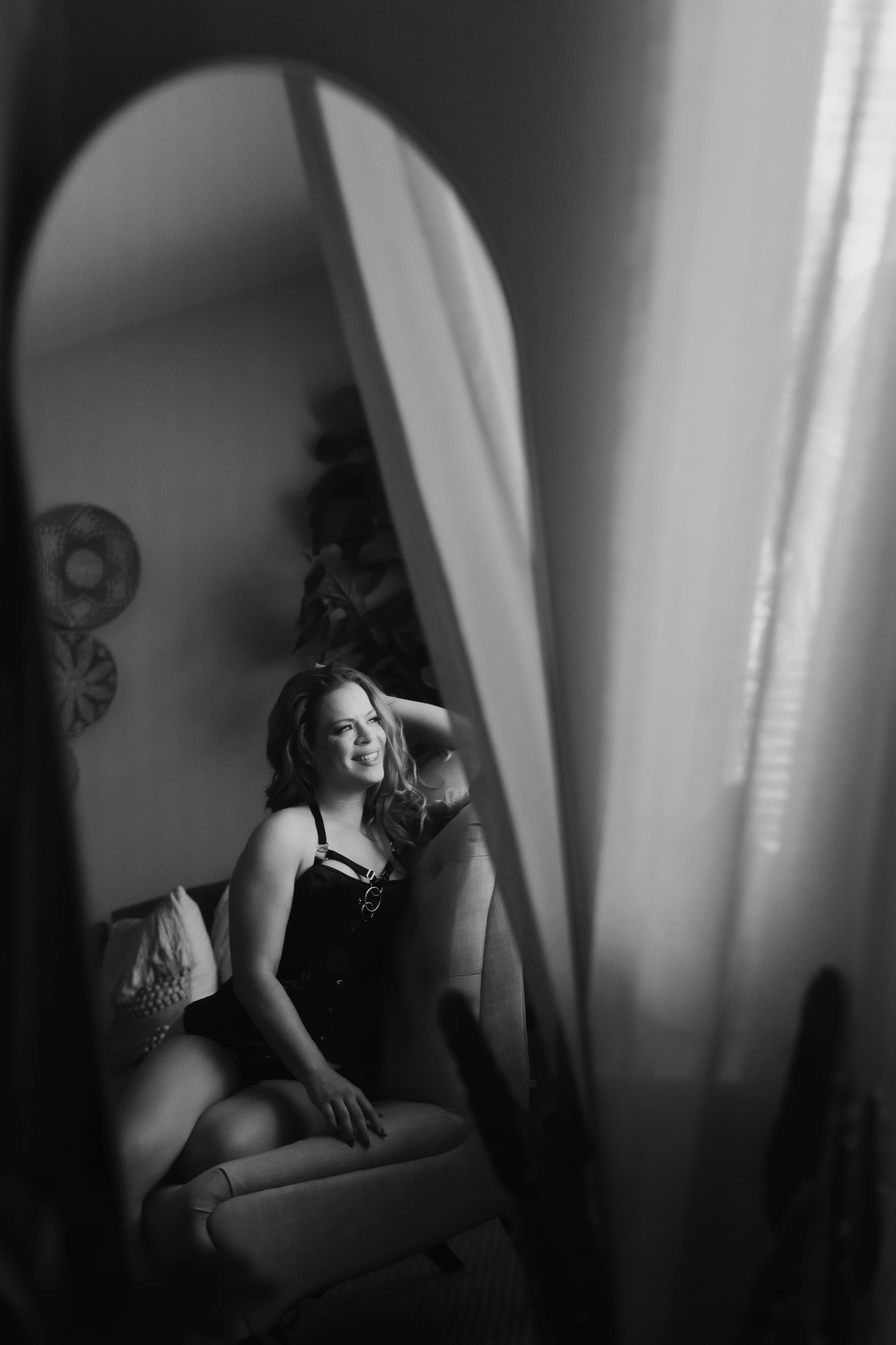 A woman with long hair sitting on a couch, smiling and wearing a black halter dress, with her knee bent and her hand resting on her leg. She is looking away from the camera, in a room with decorative wall art and a fan in the background. The photo is