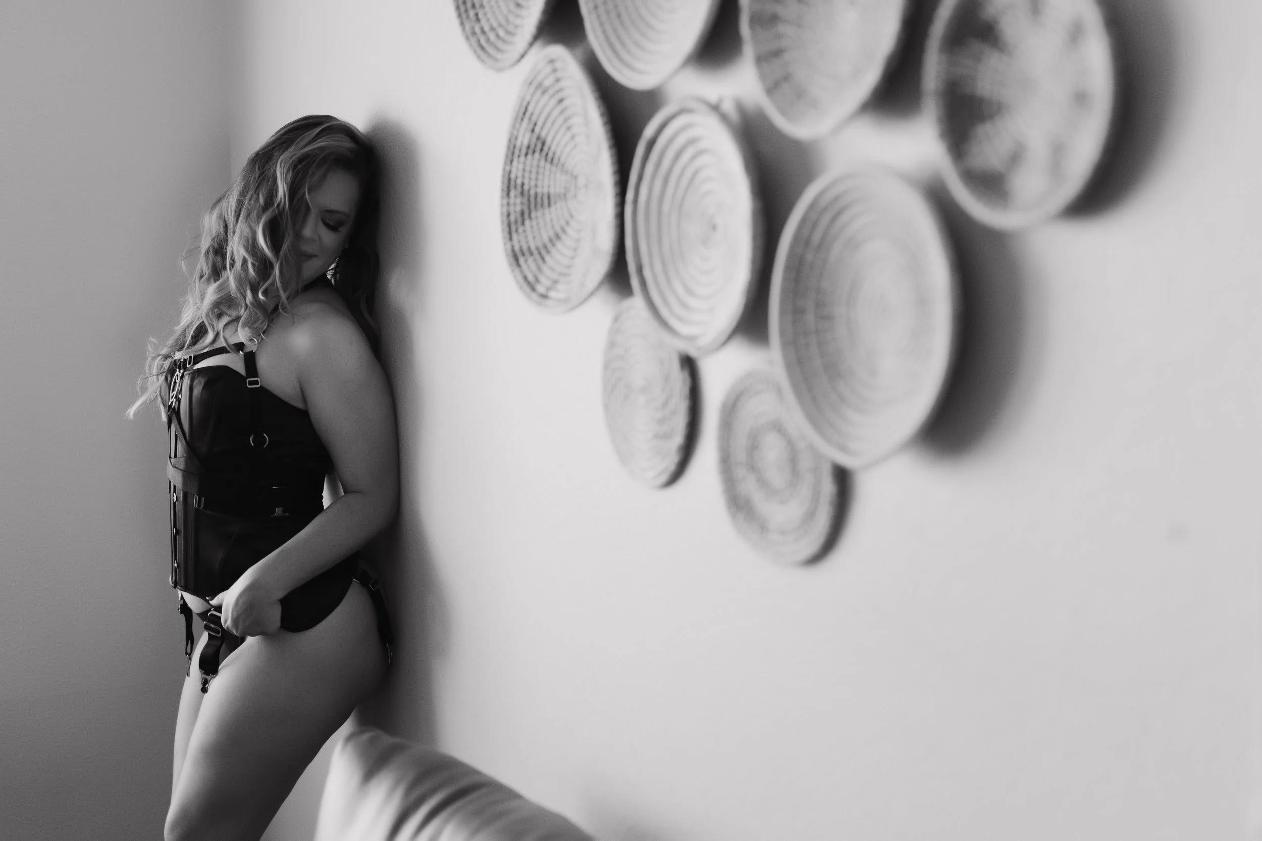 Black and white photo of a woman with curly hair wearing lingerie and a harness, standing against a wall decorated with circular wooden art.