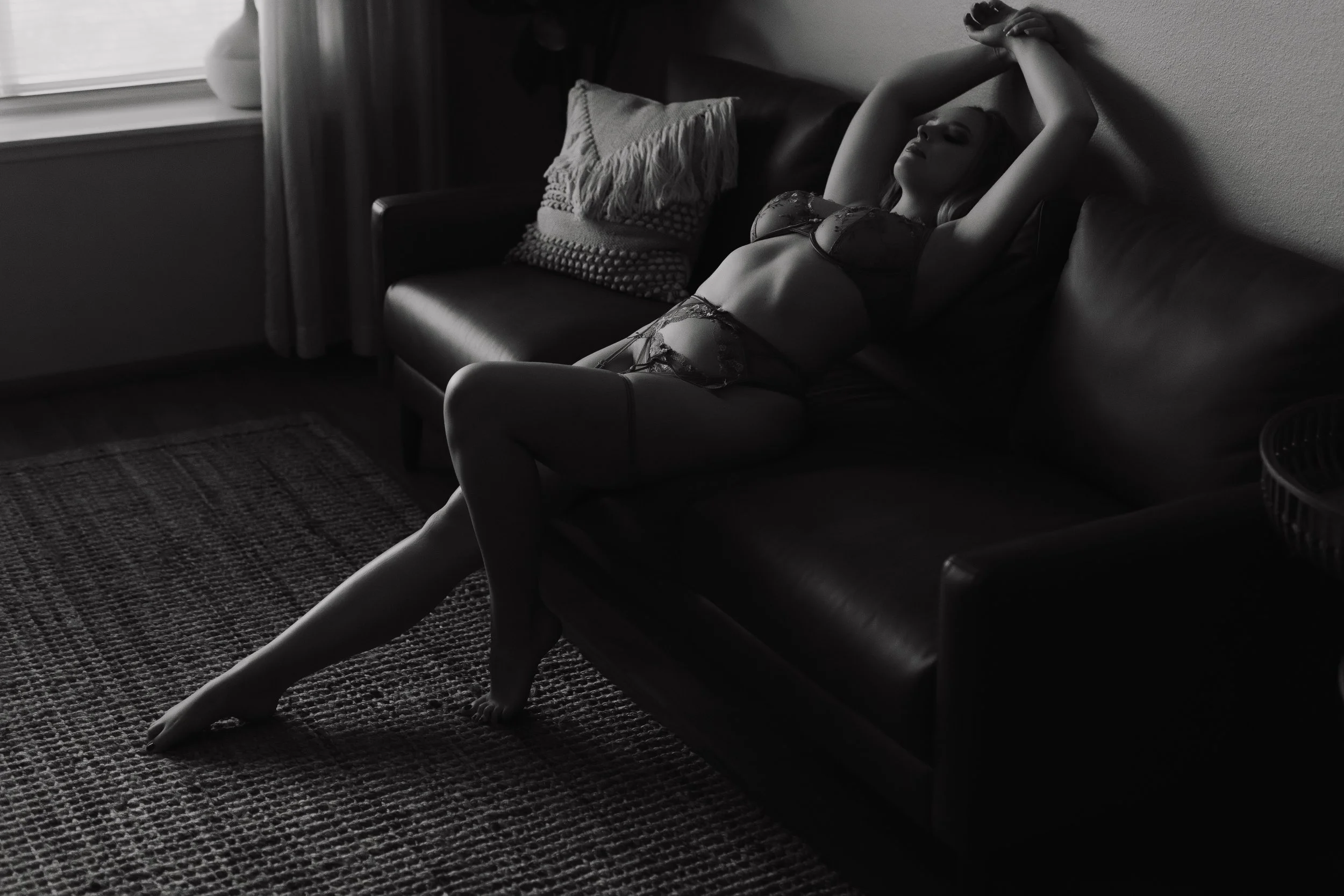 A woman wearing lace lingerie lounging on a leather couch, stretching with her arms above her head in a dimly lit living room.