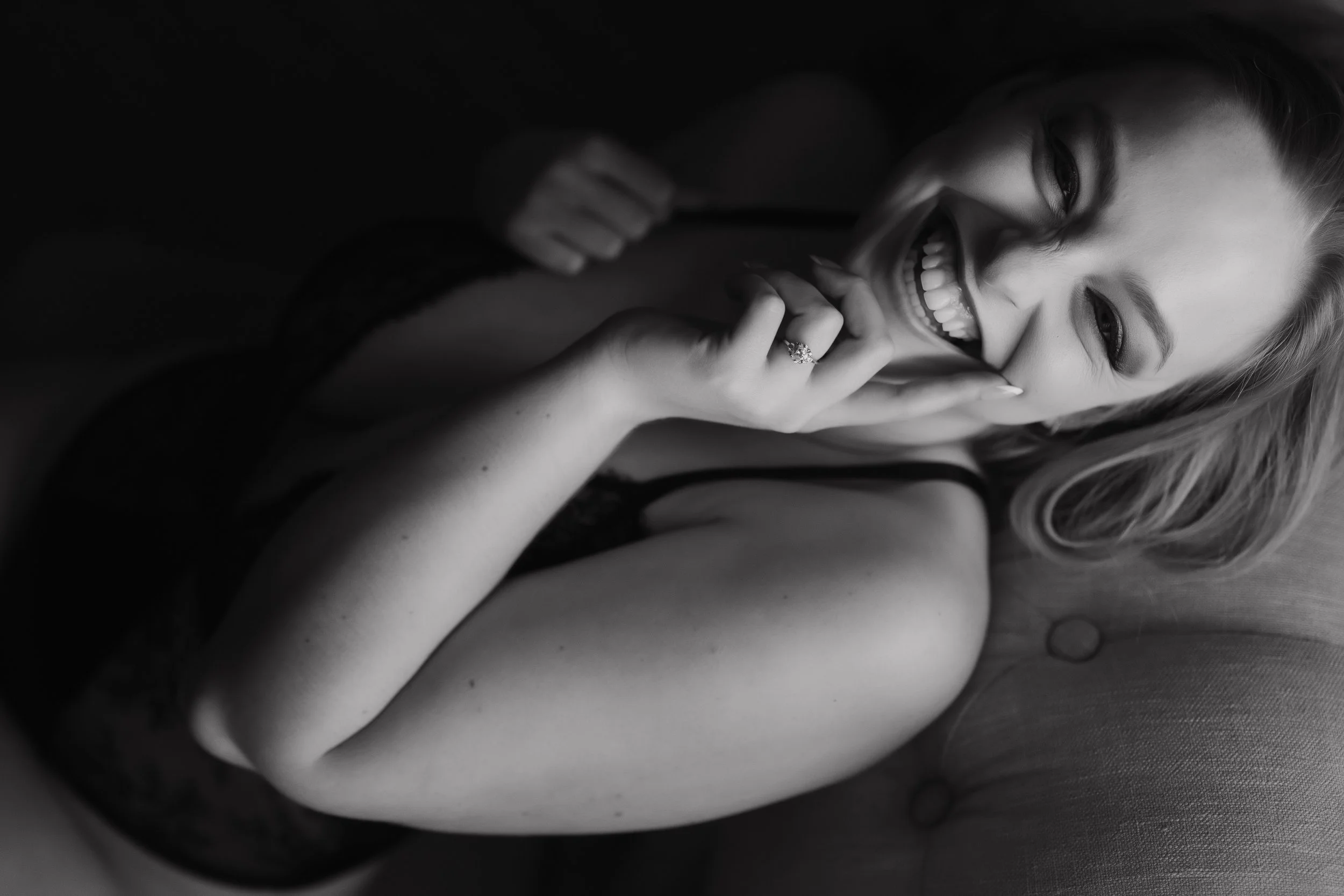 A woman with light hair smiling and biting her finger, wearing a ring and dark clothing, in a black and white portrait.