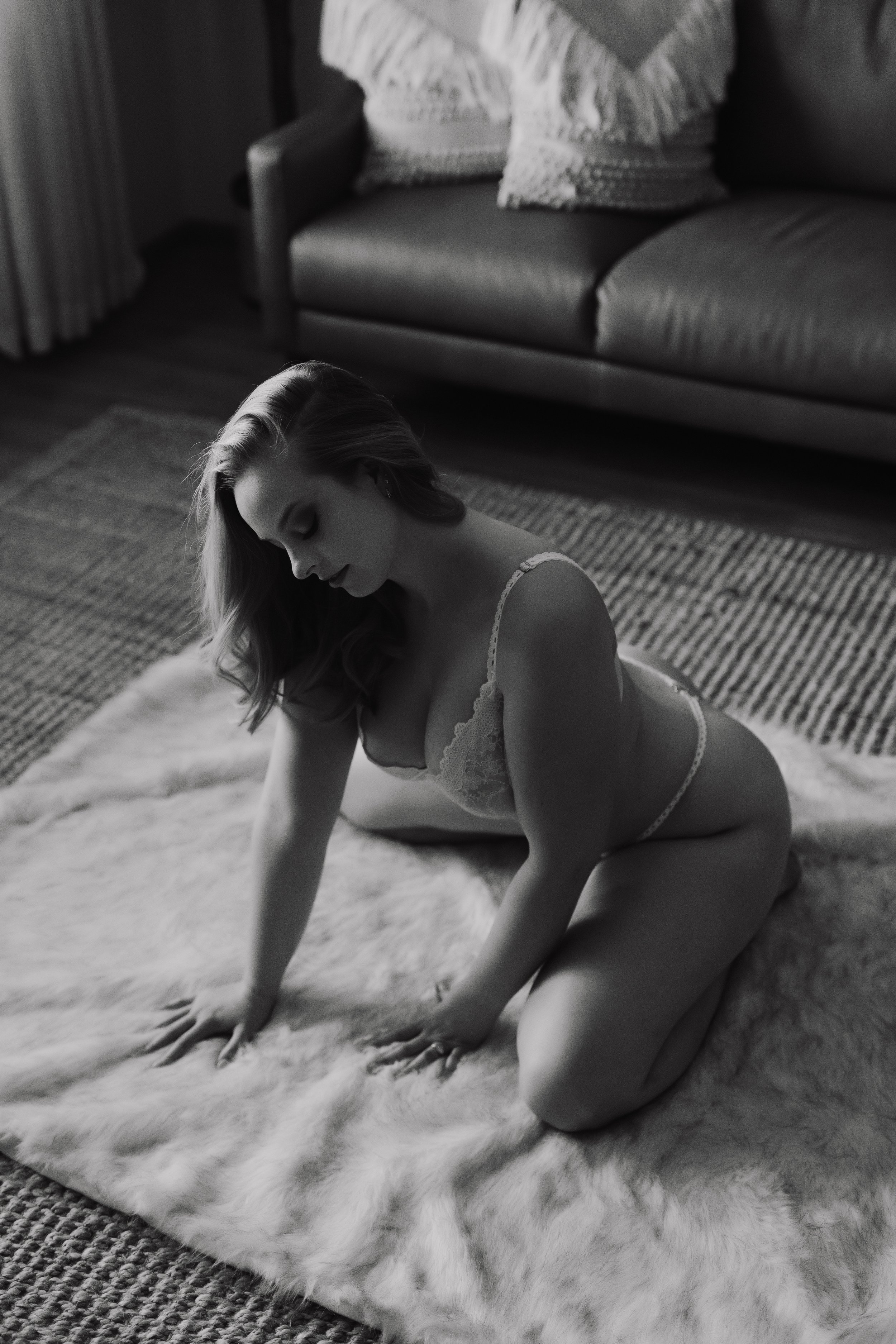 A woman with long hair, wearing lace lingerie, kneeling on a fluffy rug on the floor in a living room setting. She appears to be looking down with a calm expression. In the background, there is a sofa with pillows and curtains, and a measurement rug 