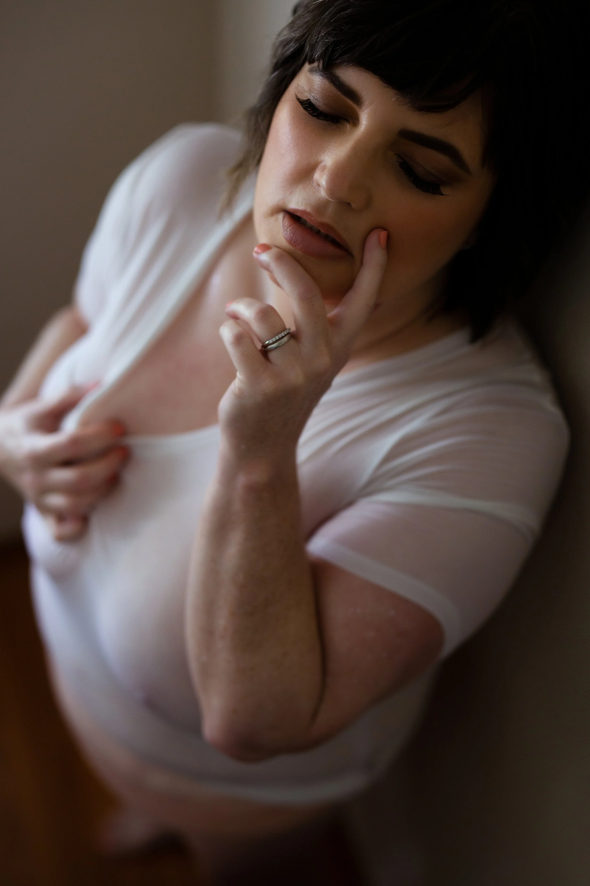 A woman with short dark hair, makeup, and a ring on her finger, wearing a white t-shirt, touching her lips with her hand, and tilting her head back.