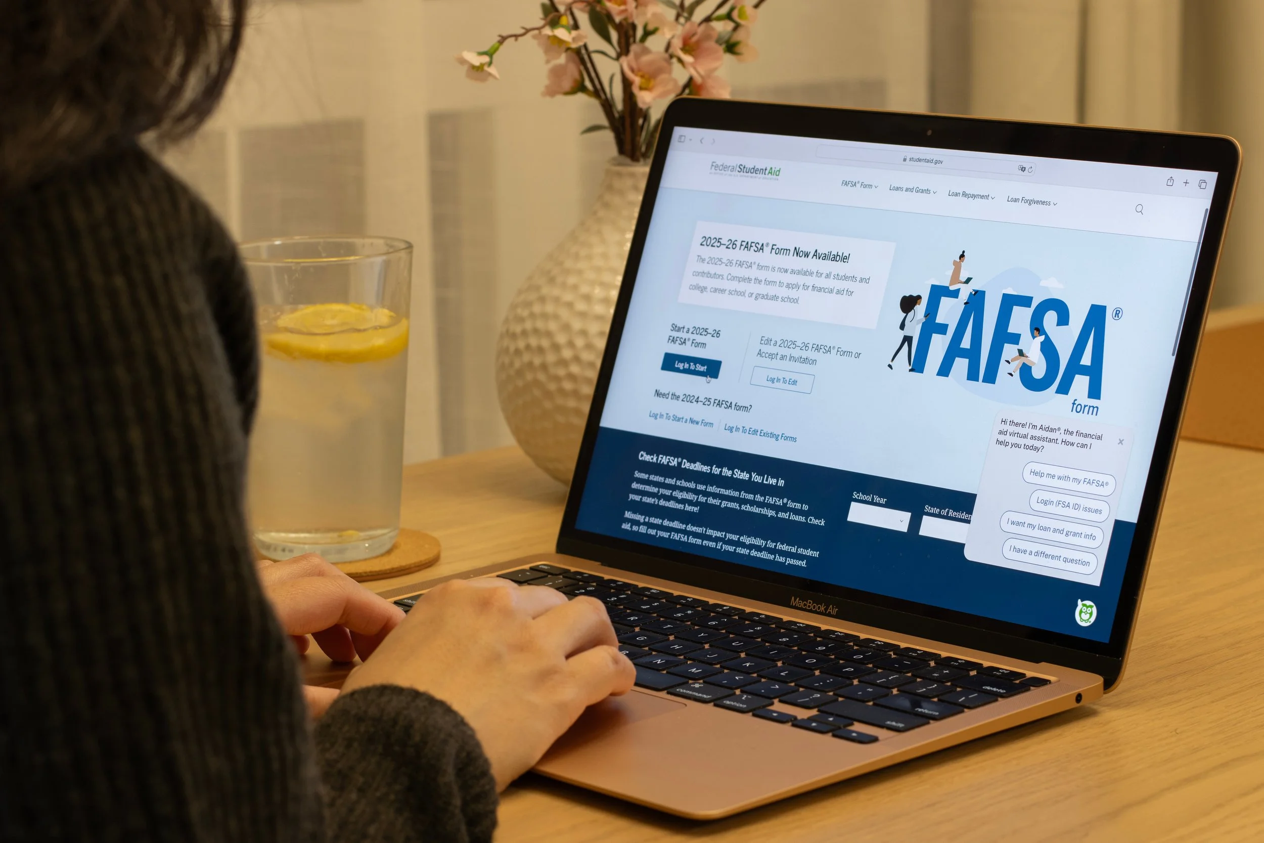 Two women sitting on a patterned couch looking at a laptop. The woman on the left is wearing an orange shirt, and the woman on the right is wearing a pink sweater and gold necklace.